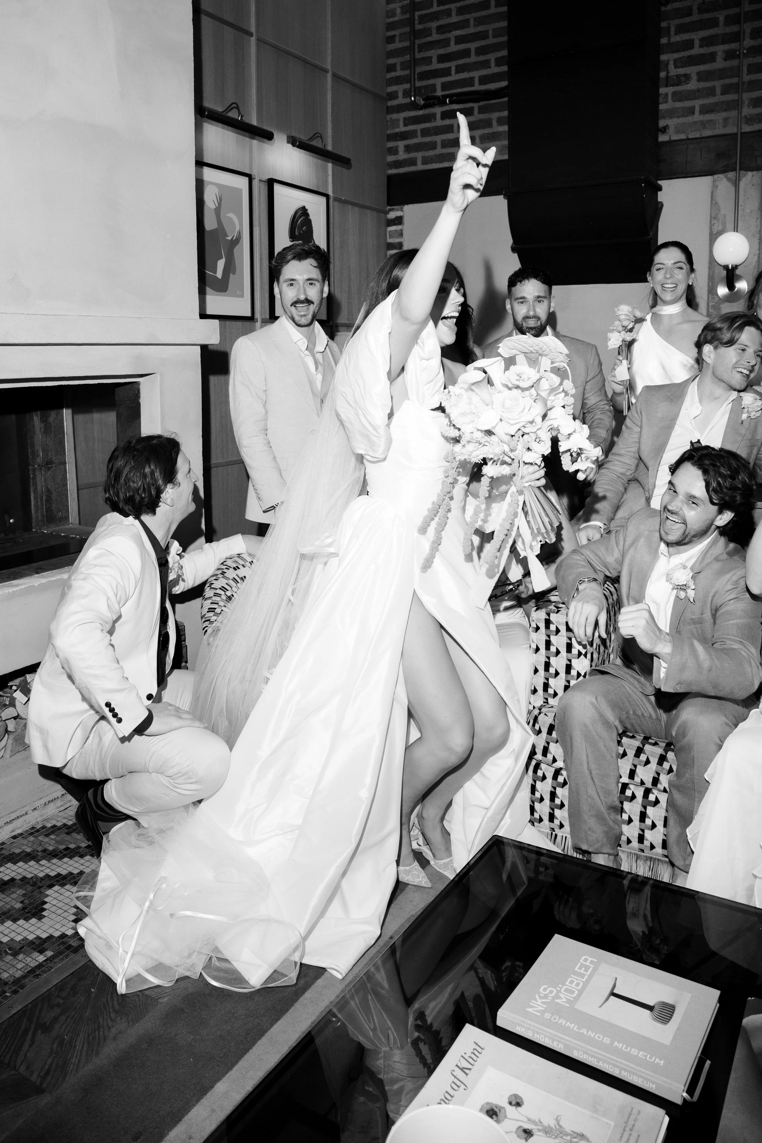 A black and white photo of a wedding celebration with a bride holding a bouquet, surrounded by guests, some seated and some standing, indoors with artwork on the walls.