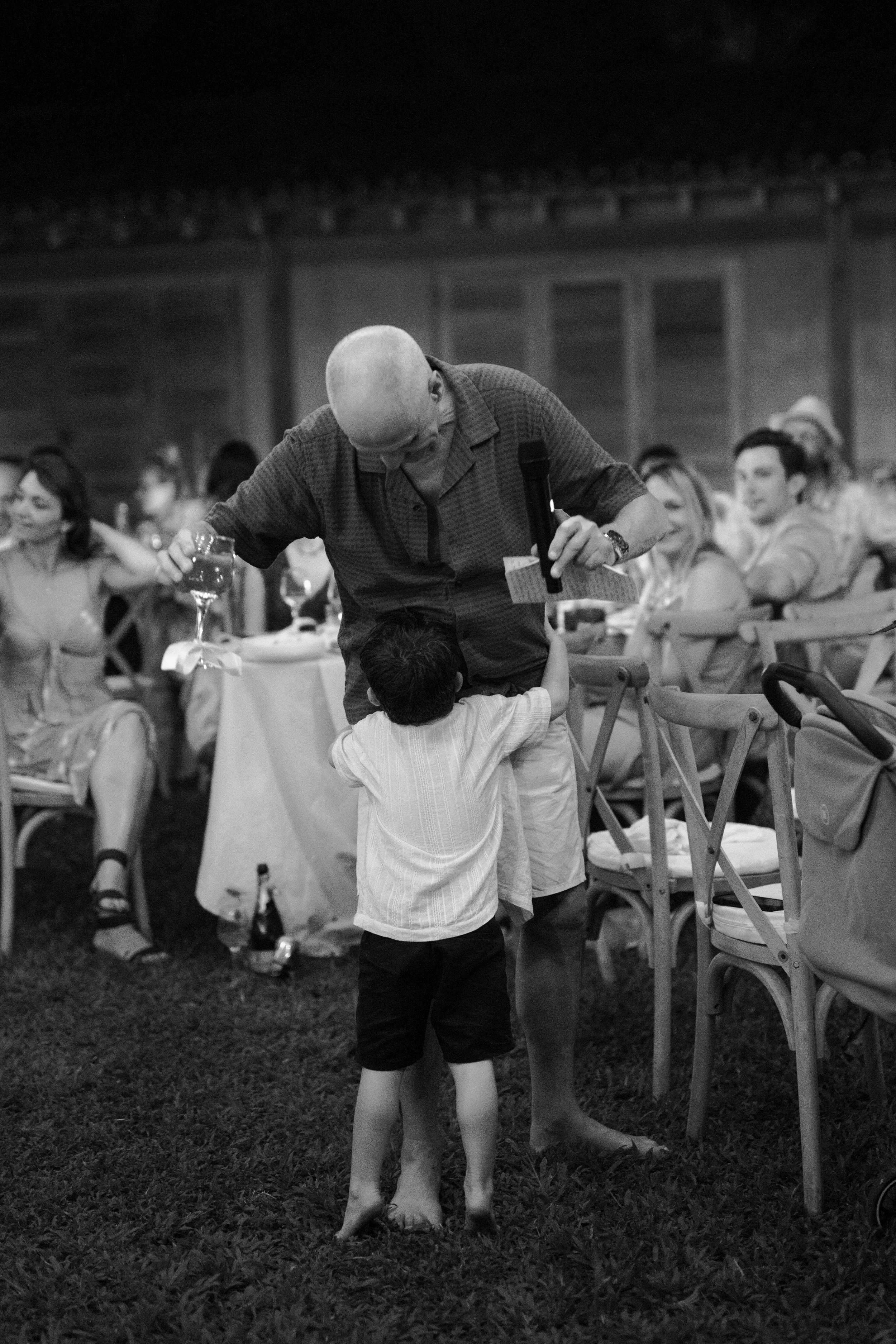 A man and a child hugging at a social gathering, with people seated at tables in the background.
