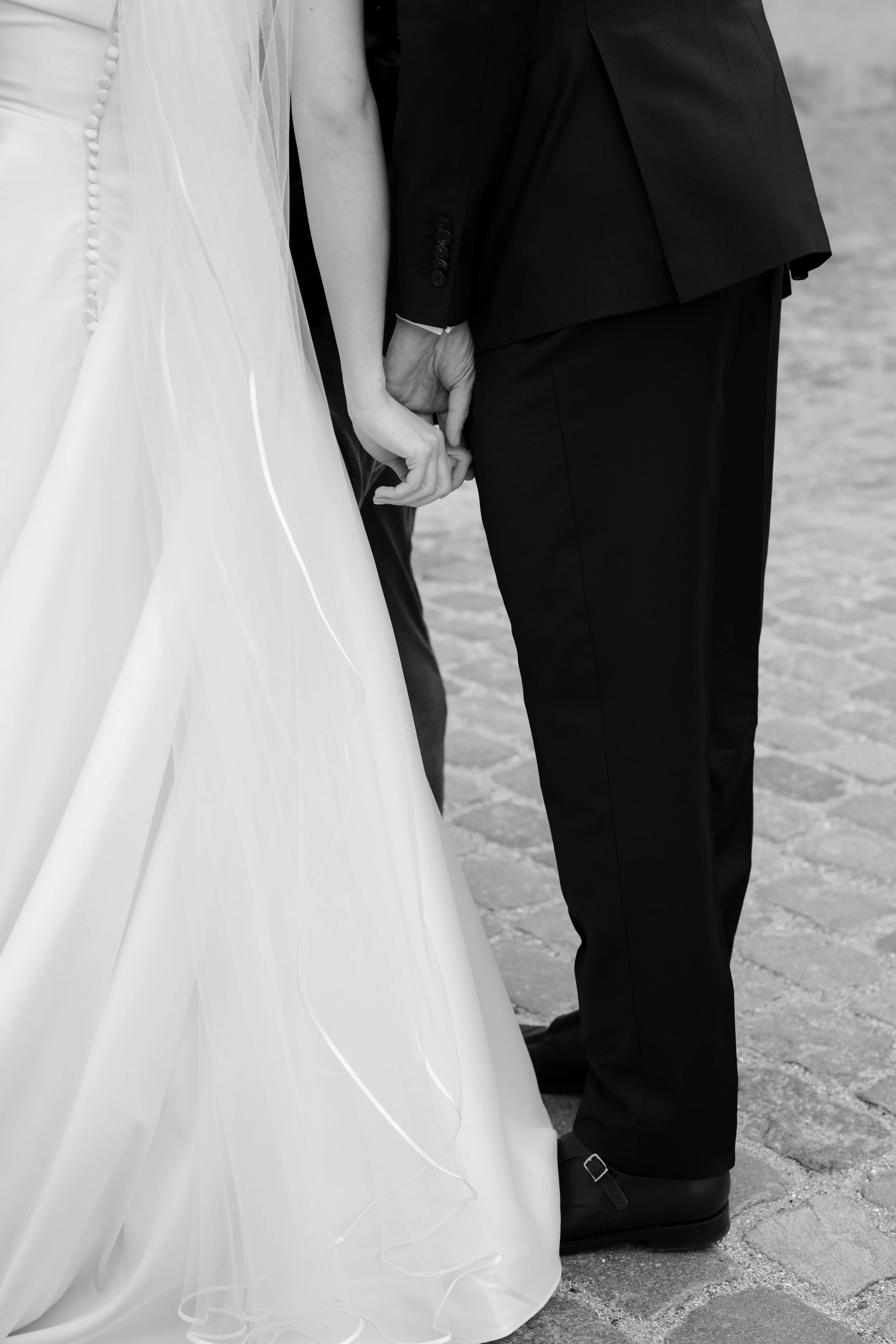 Close-up of a bride and groom holding hands, standing on cobblestone ground, black and white photograph.