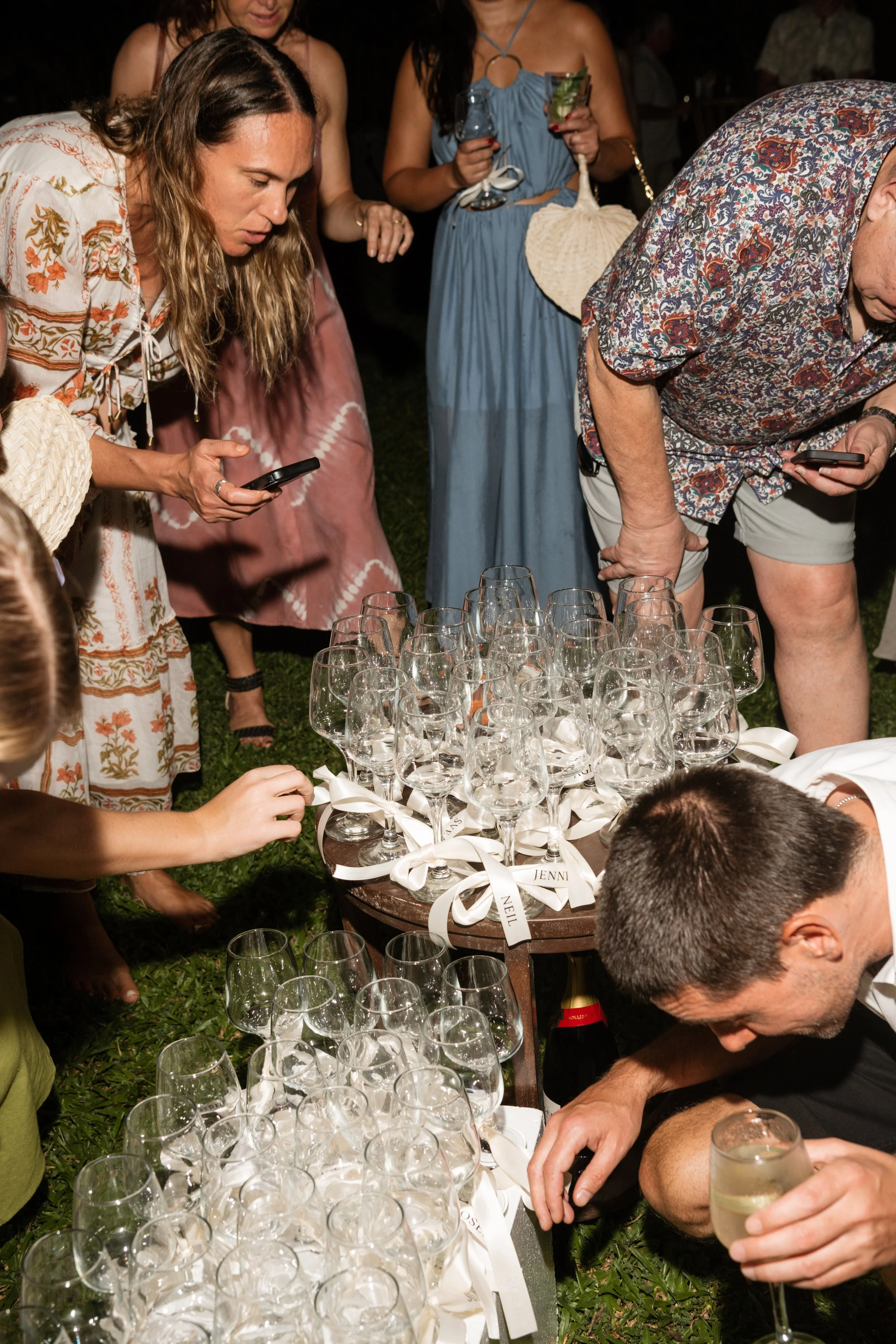 People gathered outdoors, decorating glasses on a table for a celebration or party at night.
