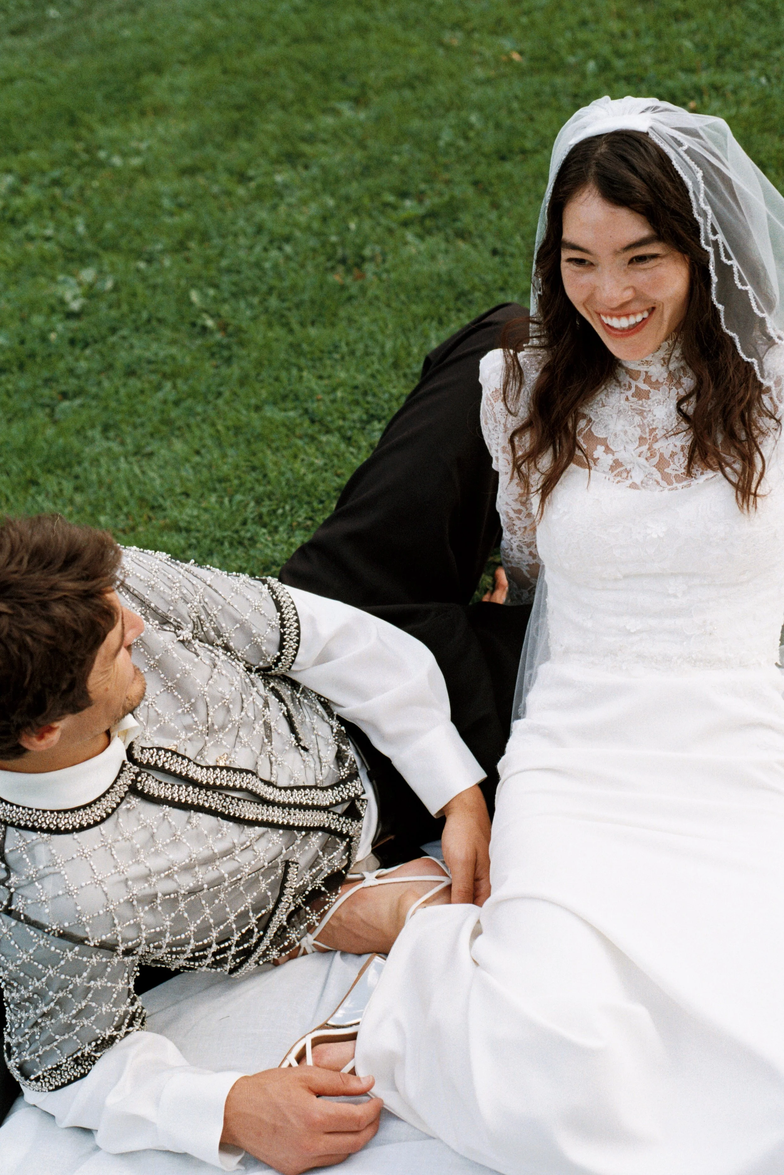 A bride and groom lying on grass, smiling and holding hands, during their wedding celebration.