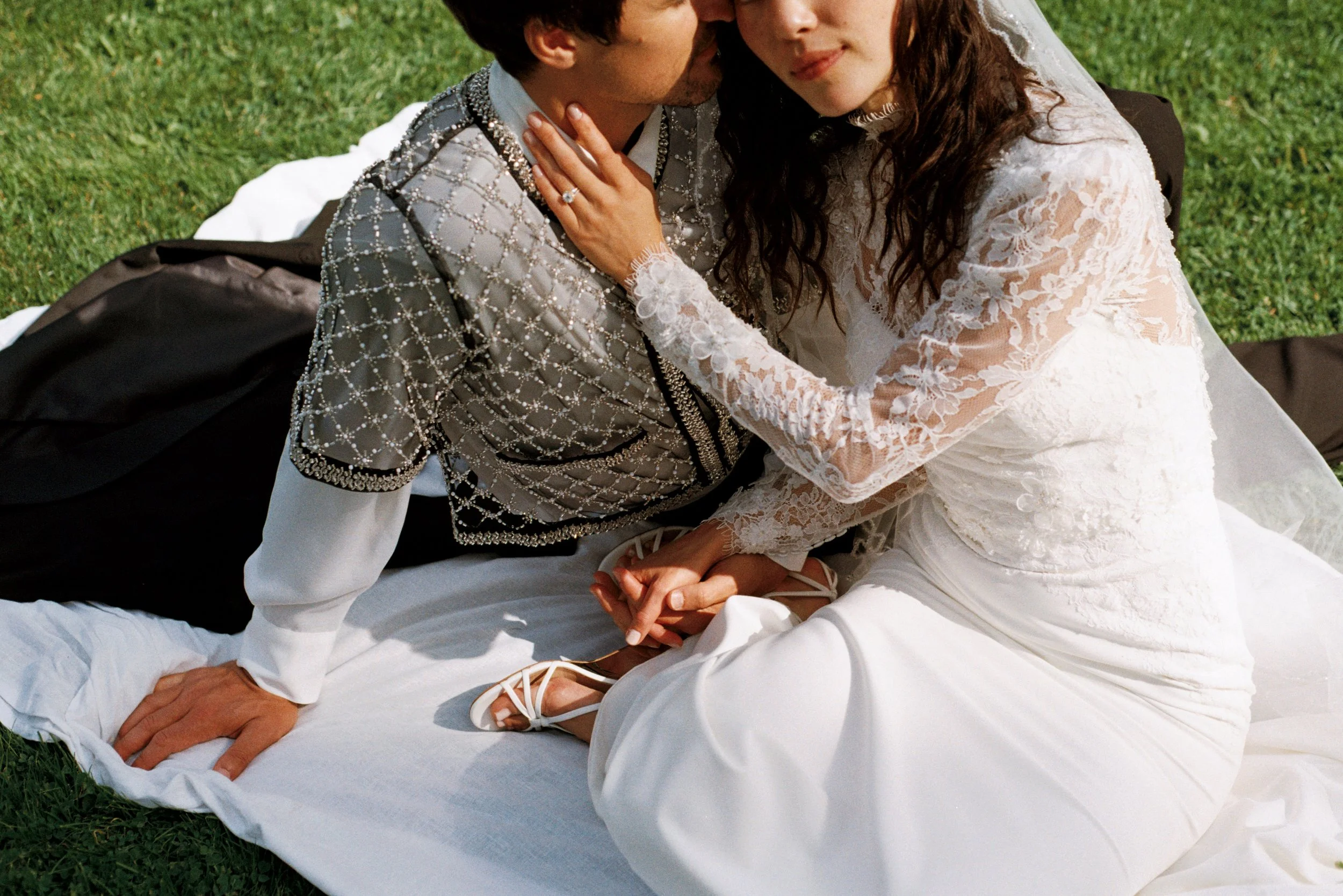 A bride in a lace wedding dress sits on a white blanket on the grass, holding hands with a groom in a gray patterned outfit. The groom, also dressed in elaborately decorated clothing, leans in for a kiss. The couple is outdoors with a green lawn in the background.