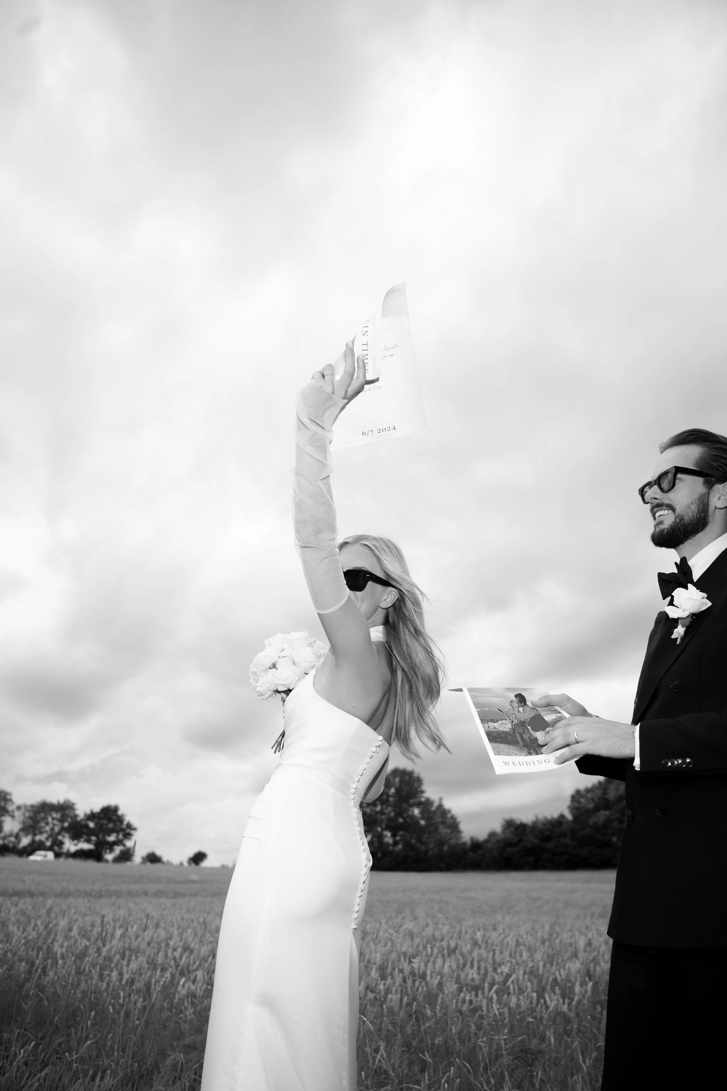 A bride and groom outdoors, with the bride raising a wedding program in the air and the groom holding a wedding photo, both smiling.