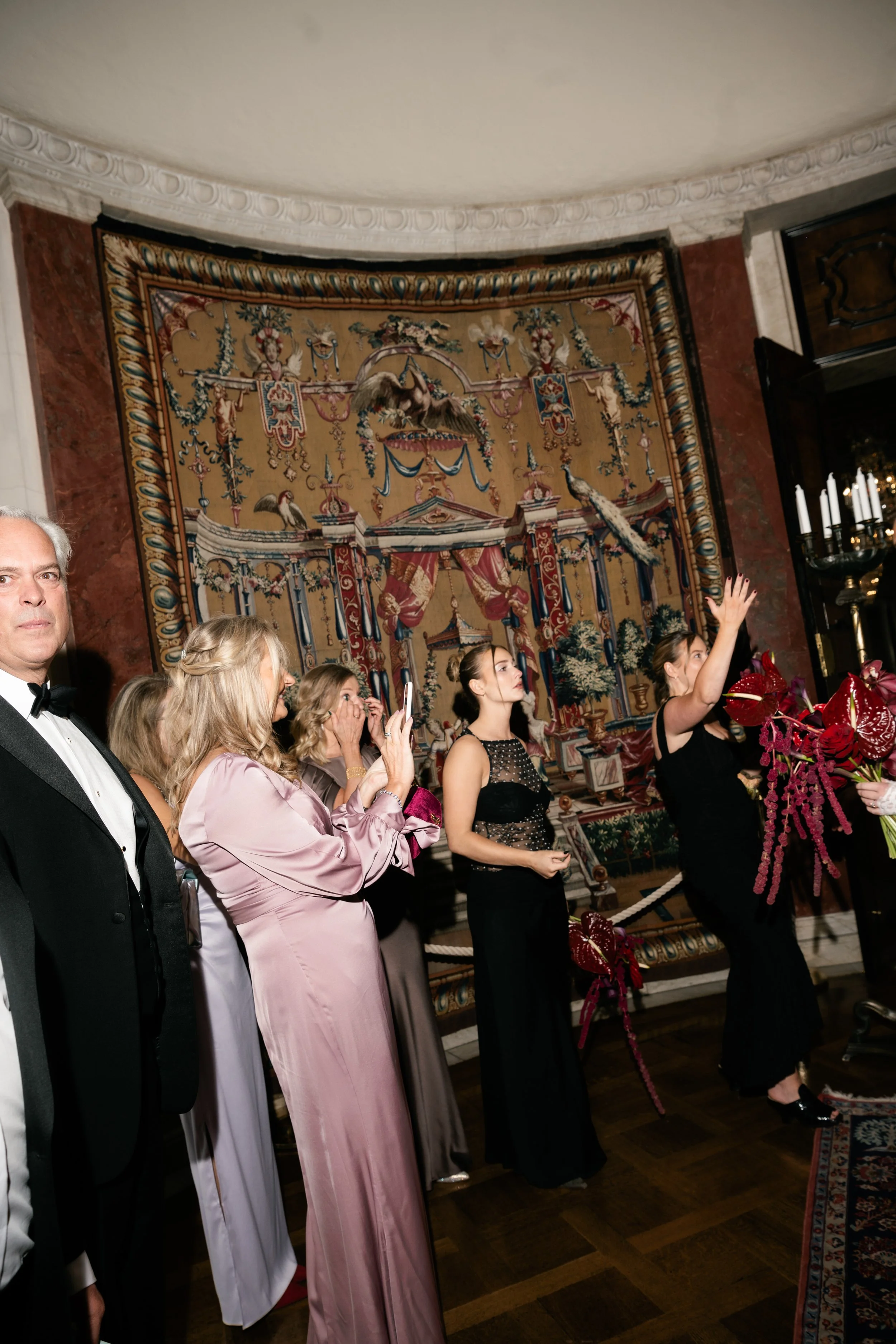 A group of people dressed in formal attire standing in front of a tapestry at a reception or celebration. One woman is raising her hand, while others are taking photos or watching.