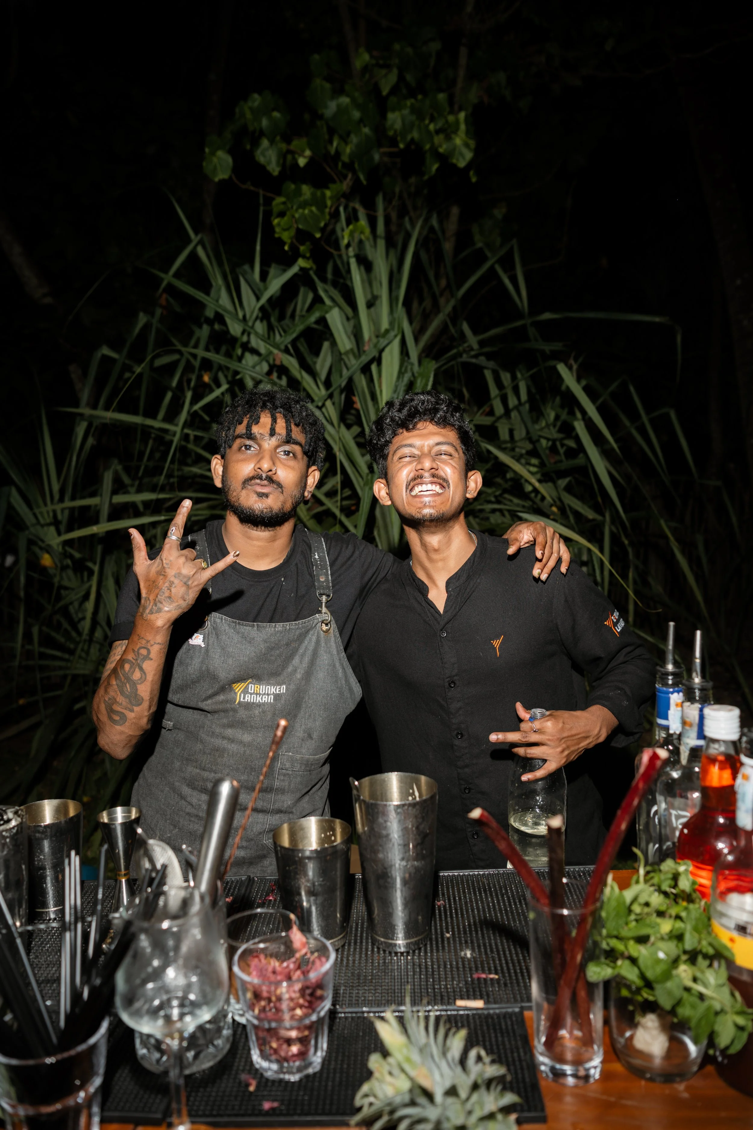 Two men behind a bar with cocktail tools and bottles, one making a hand gesture, the other smiling, outdoors at night with plants in the background.