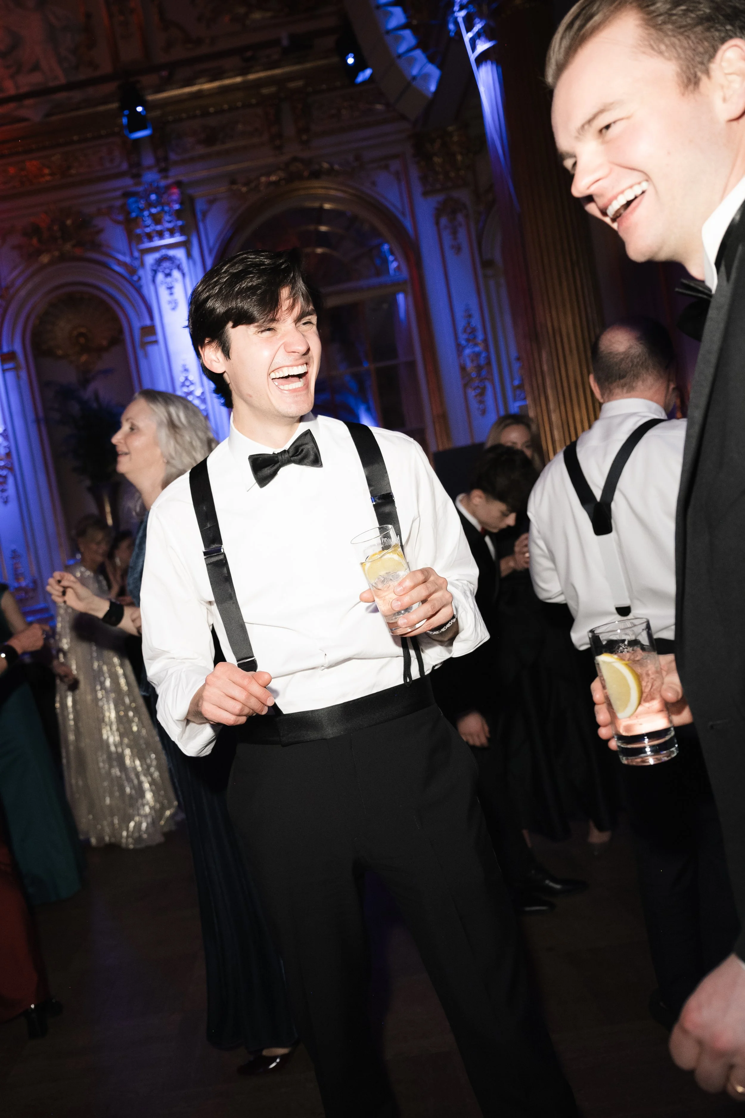 People dancing and socializing at a formal event in an ornate decorated ballroom, with blue lighting and large windows.