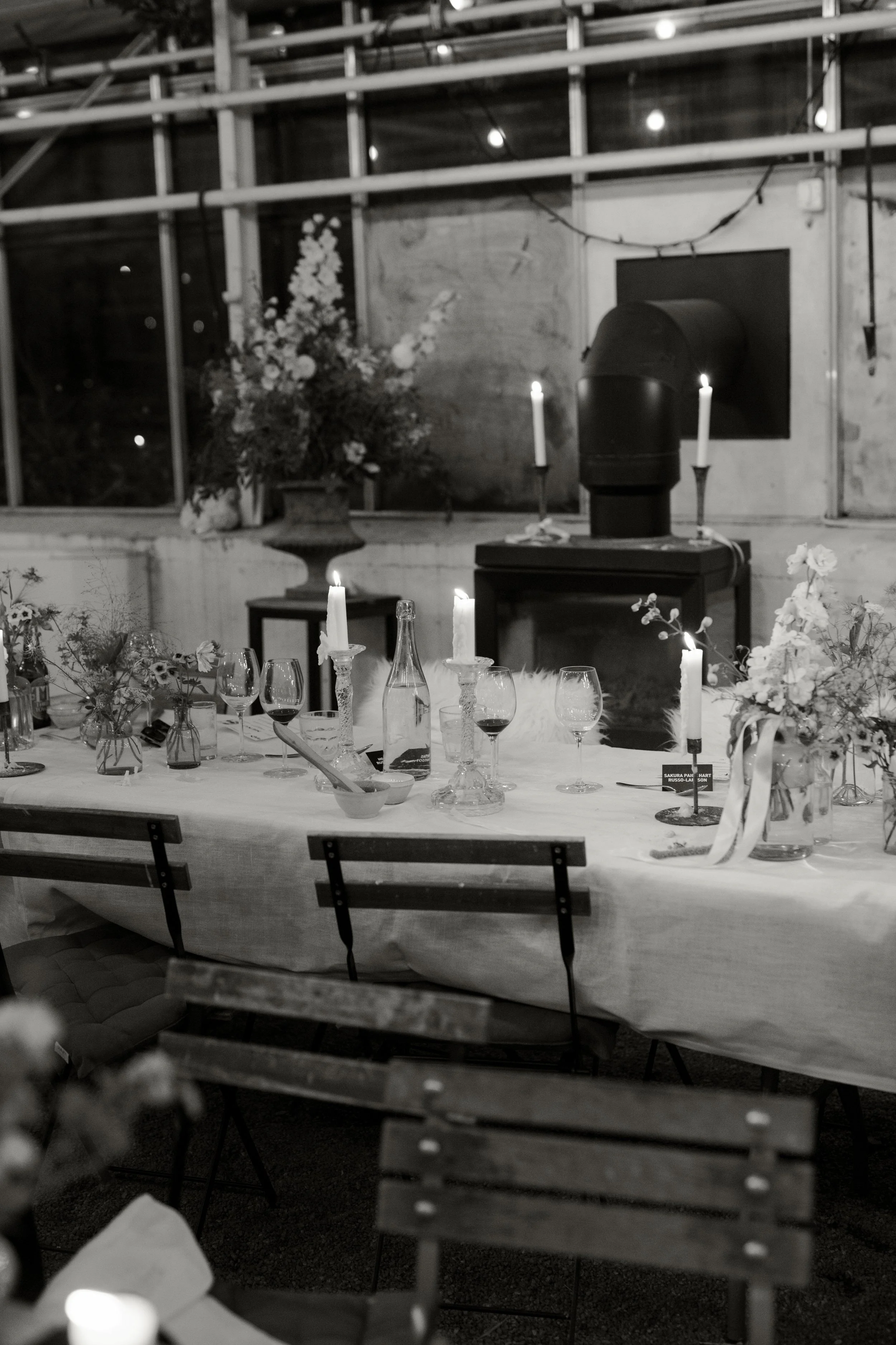 A dining table set with candles, flowers, and glasses inside a rustic room with exposed brick and large window, decorated for an event or dinner party.