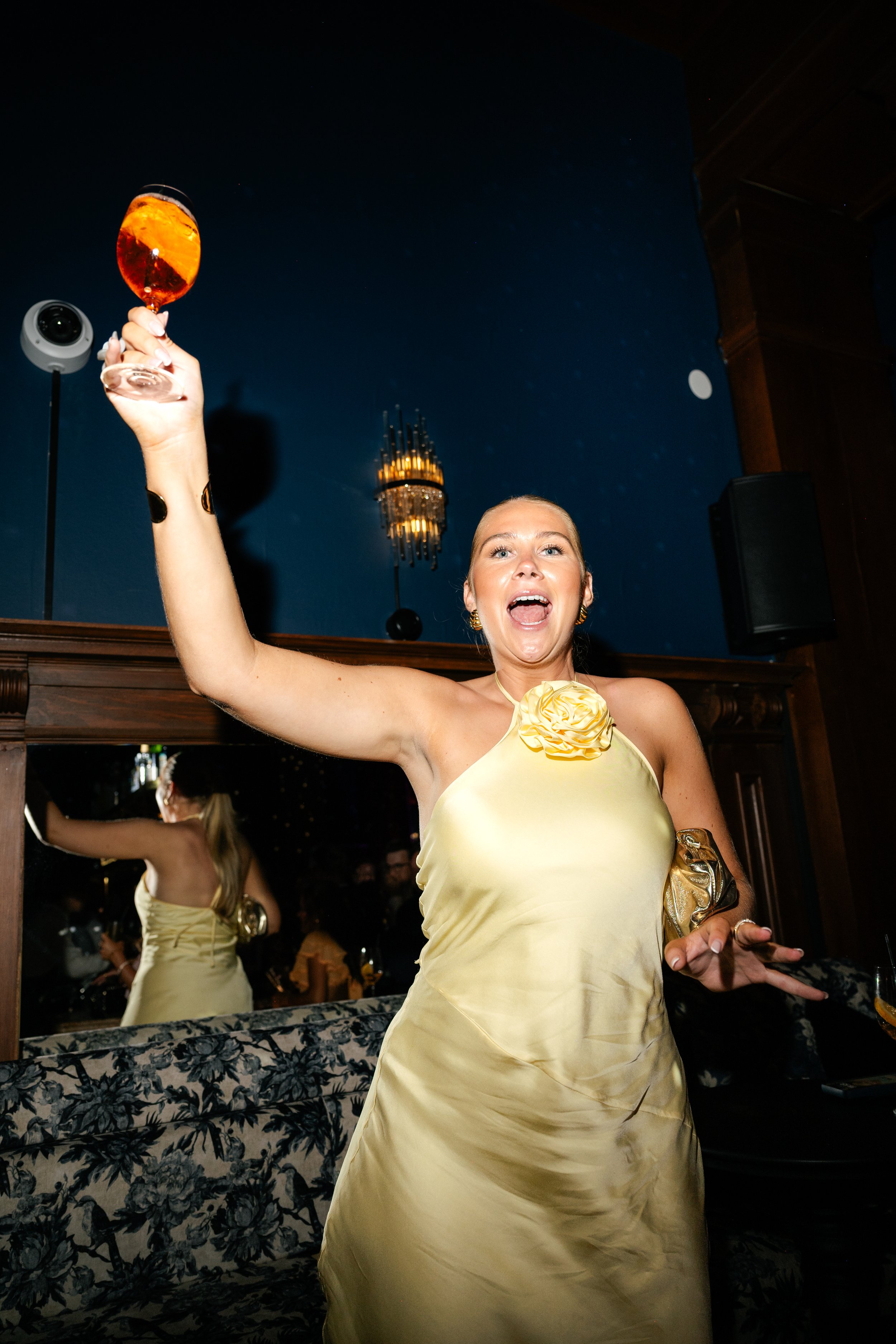 Woman in a yellow satin dress holding a glass of wine high, celebrating at a party or event.