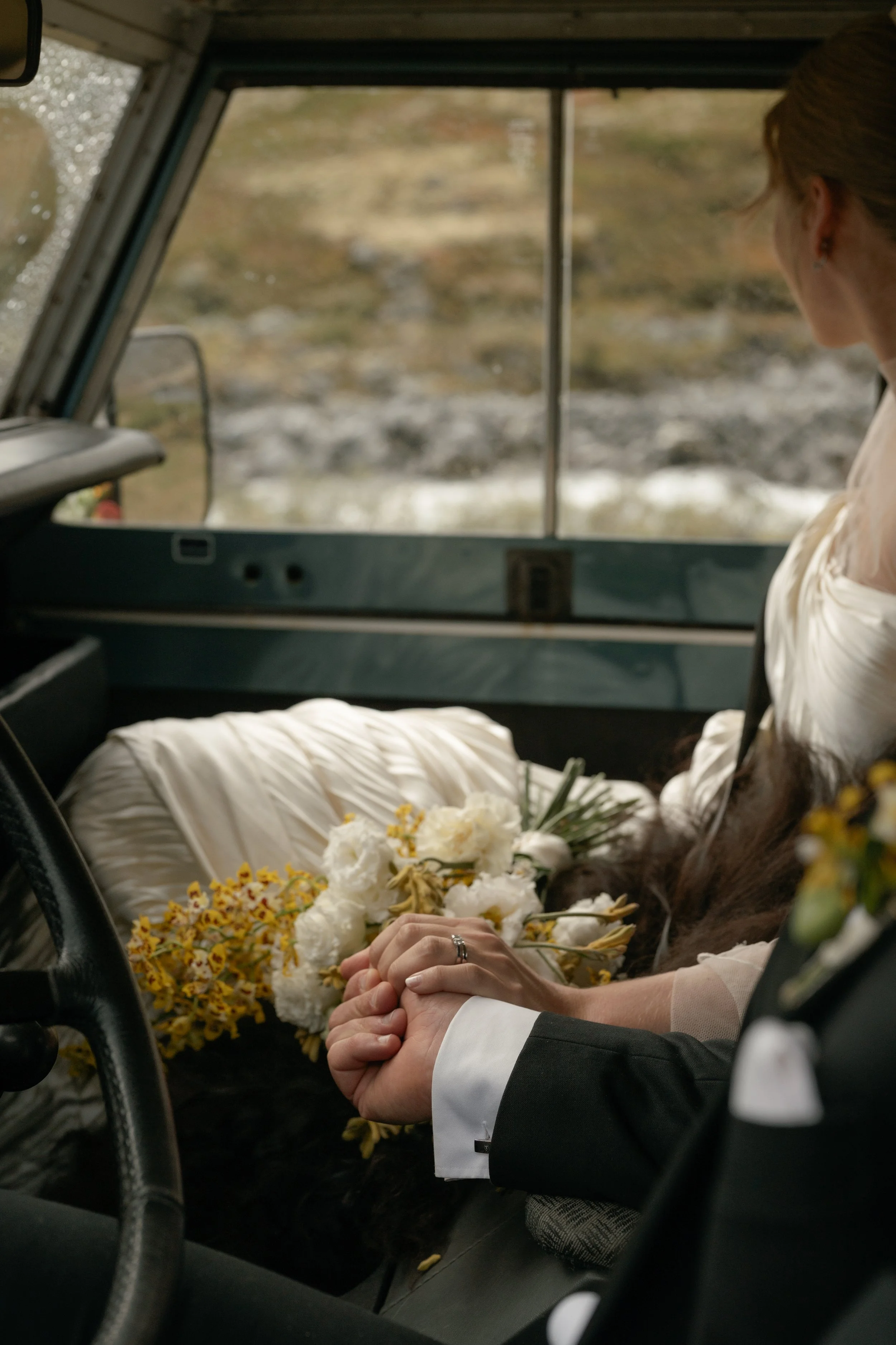 A bride and groom sitting in a vintage vehicle, holding hands, with a bouquet of white and yellow flowers on the bride's lap, during a wedding or special occasion.
