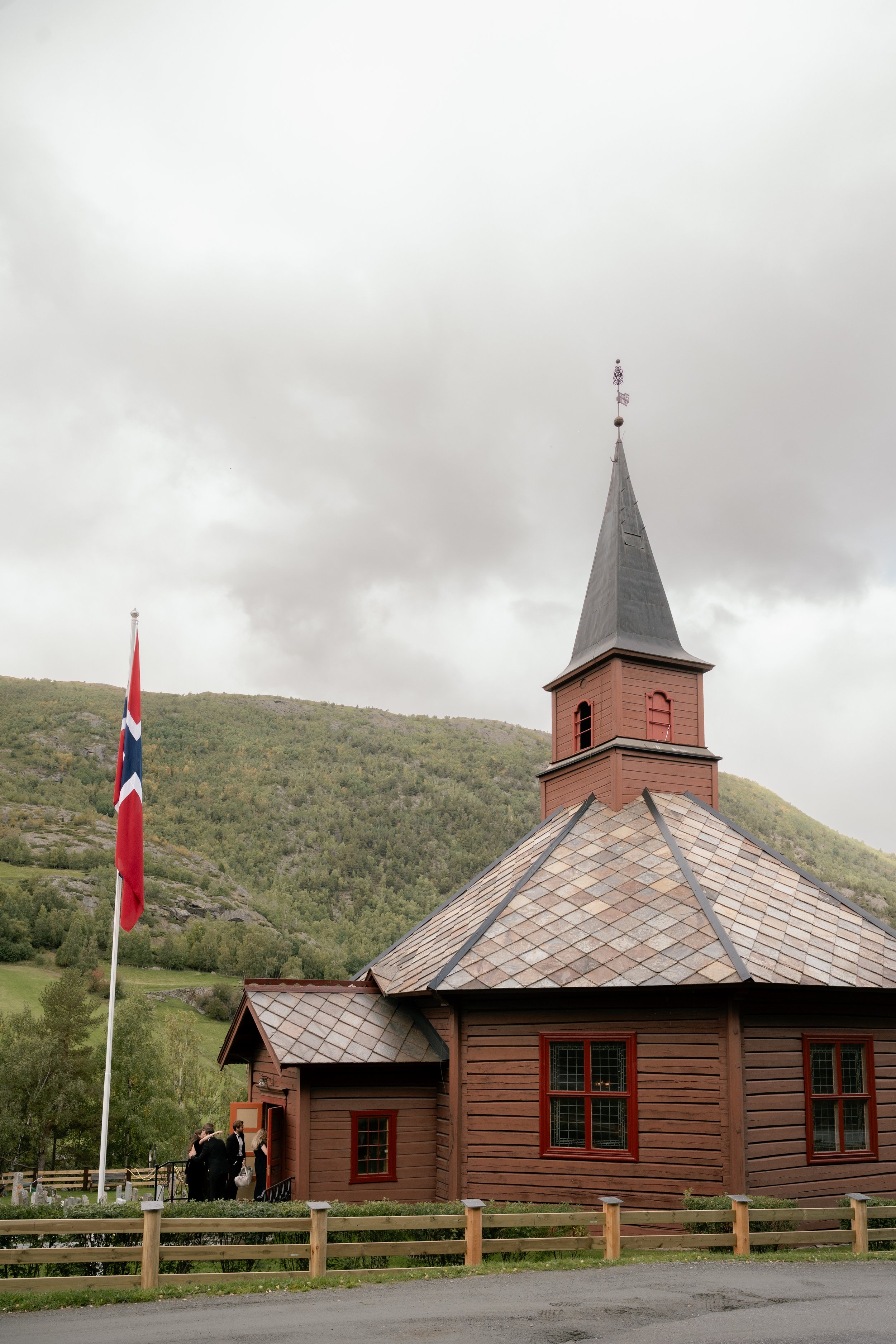A small wooden church with a red exterior, a tall steeple, and several windows, located in a green, hilly area with cloudy skies. There is a Norwegian flag on a flagpole nearby, and a group of people walking toward the church.