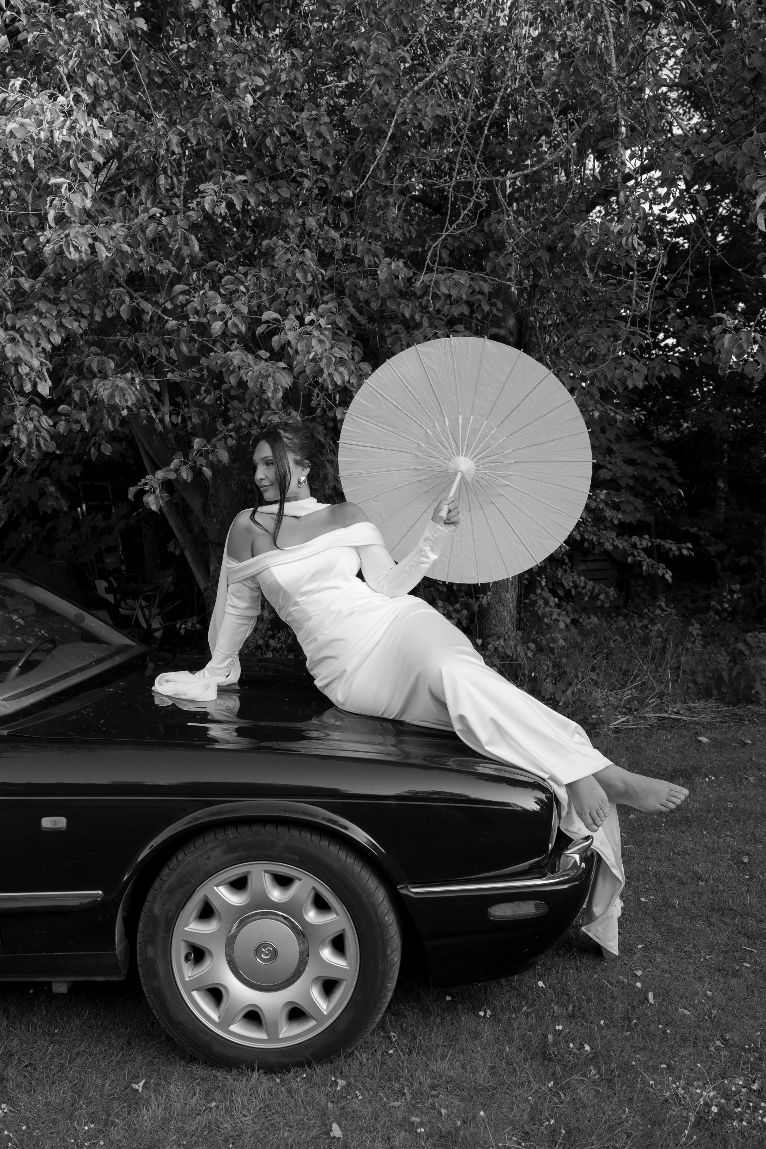 A woman in a long white dress is sitting on the hood of a black car, holding a parasol, with trees in the background.