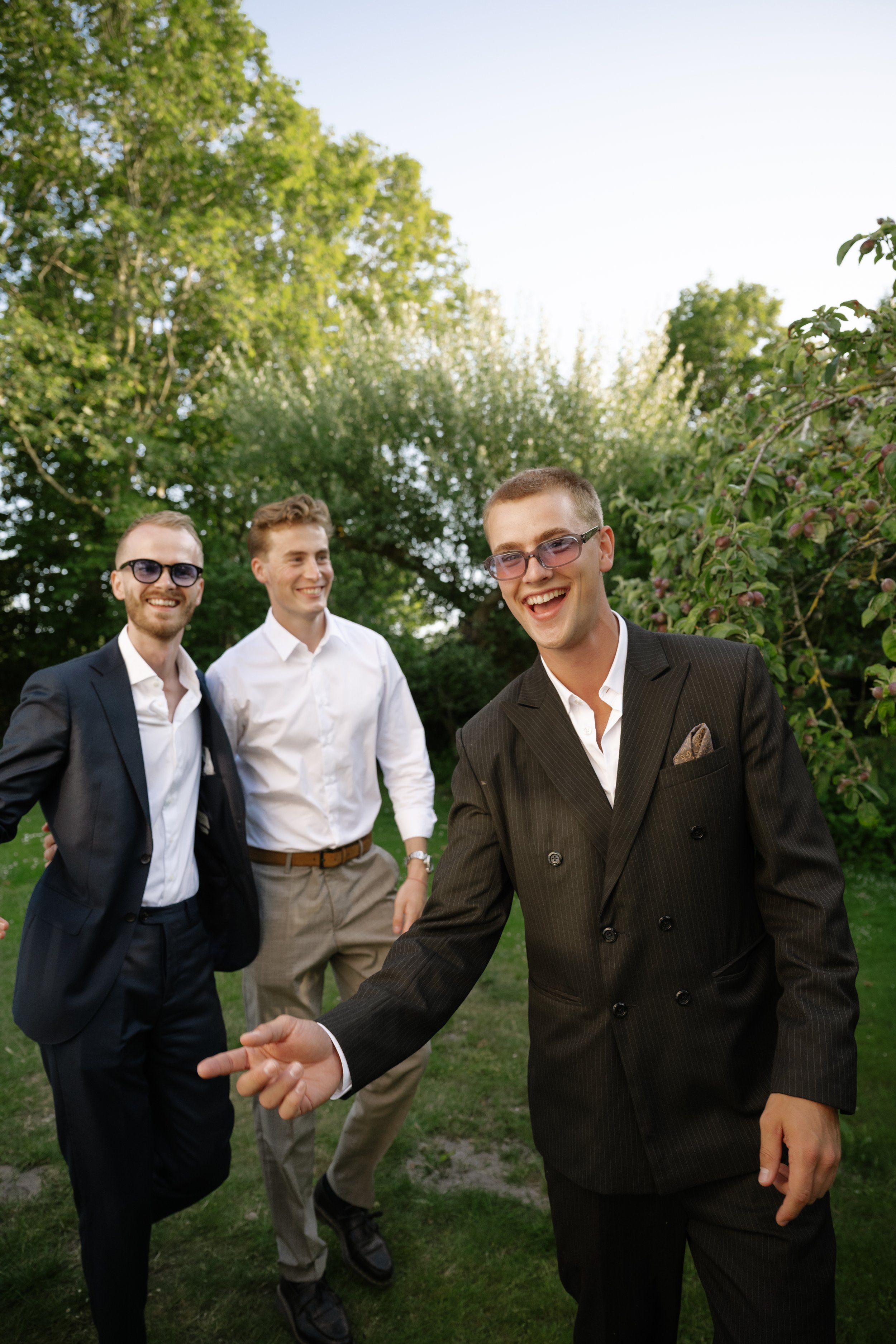 Three young men in suits smiling and enjoying themselves outdoors in a green park during daytime.