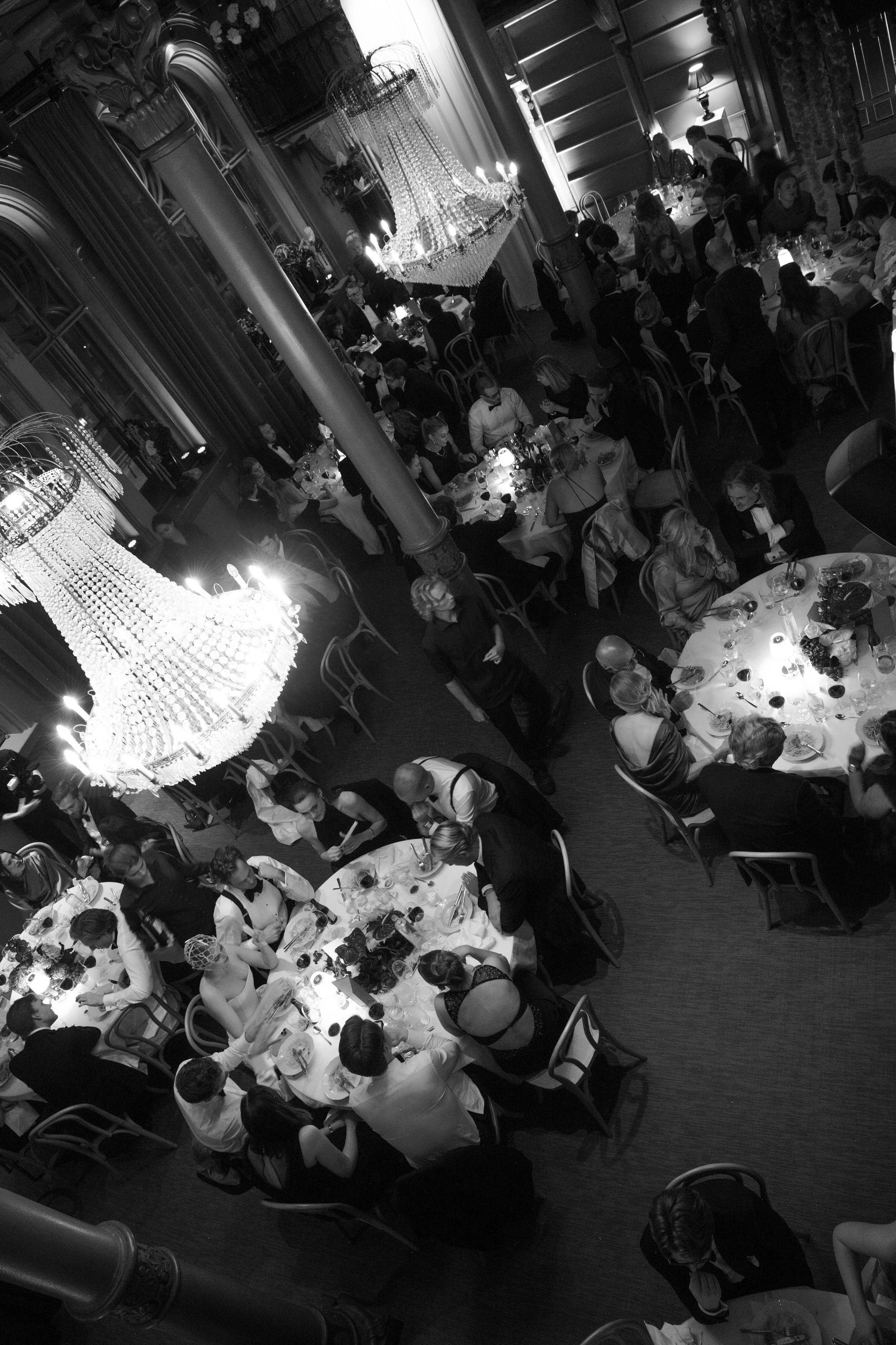 A black and white photo of a banquet hall filled with people sitting at round tables, some standing and walking, with grand chandeliers hanging from the ceiling, set for a formal event or dinner.