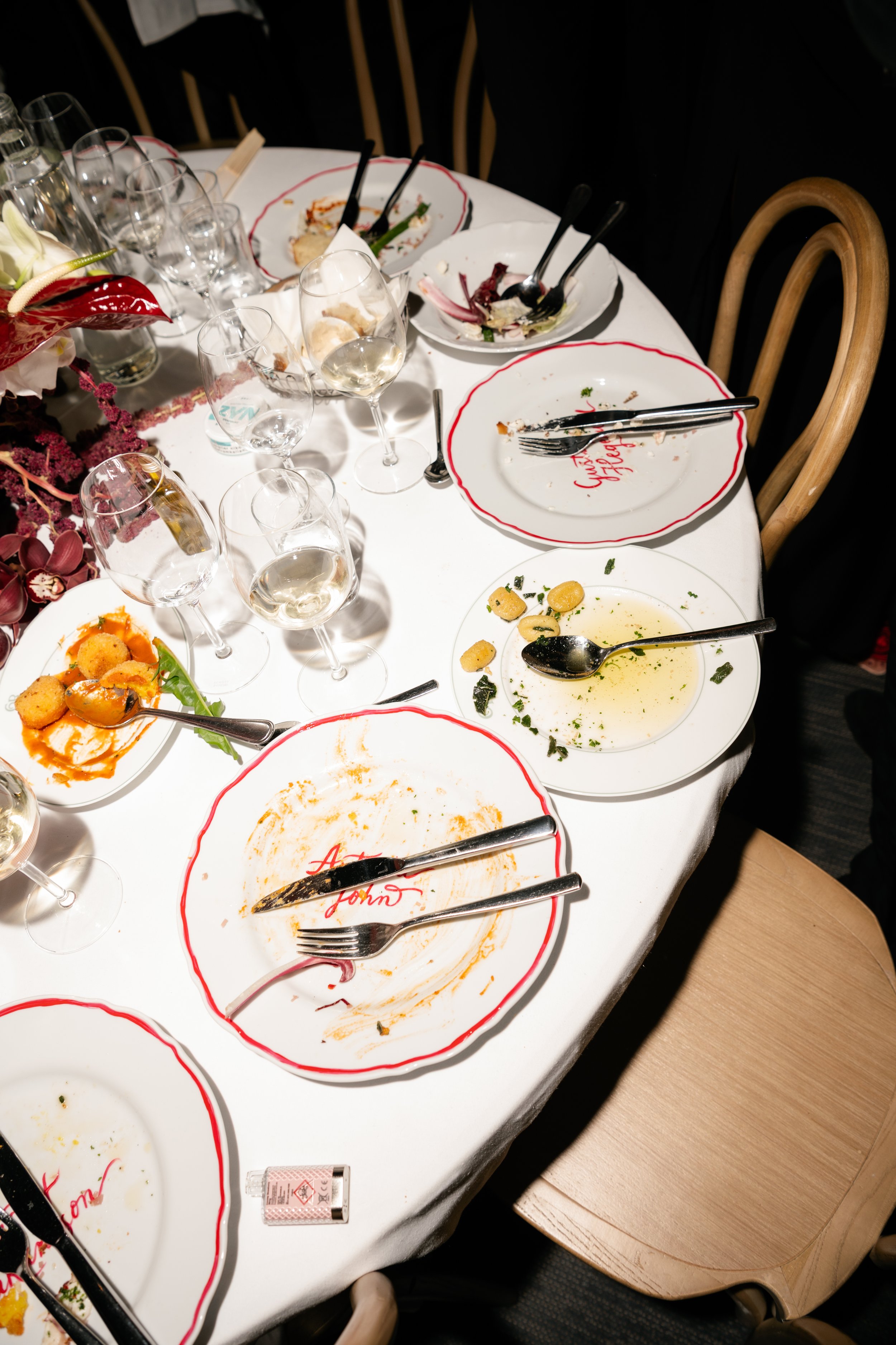 A round dining table with empty and partially eaten plates, used cutlery, several wine glasses, and some leftover food, in a dimly lit setting that suggests a finished meal at a restaurant or event.