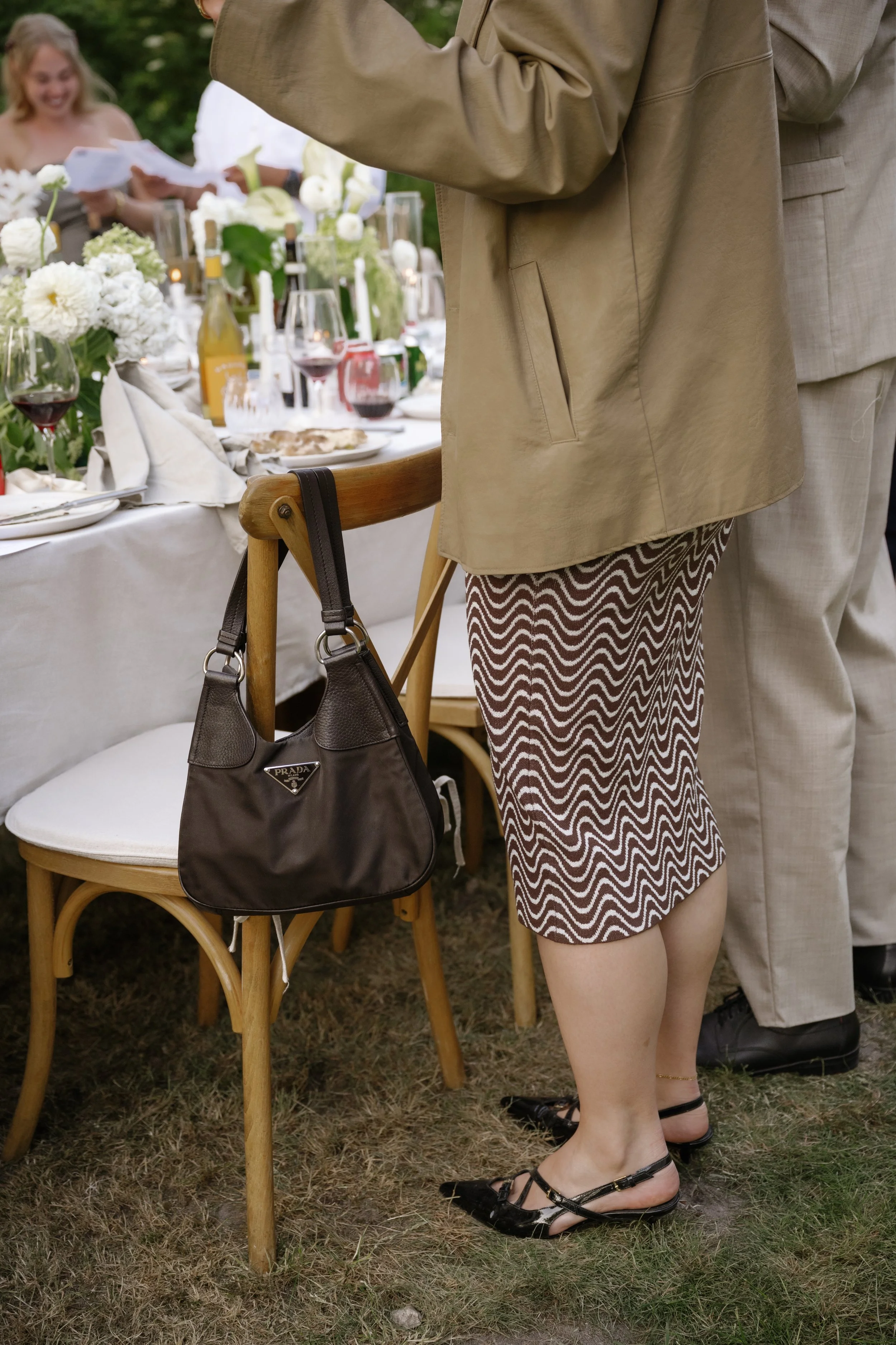Close-up of a woman at an outdoor dinner party, wearing a patterned skirt, black shoes, and a beige jacket, with a Prada handbag hanging on the back of a wooden chair.