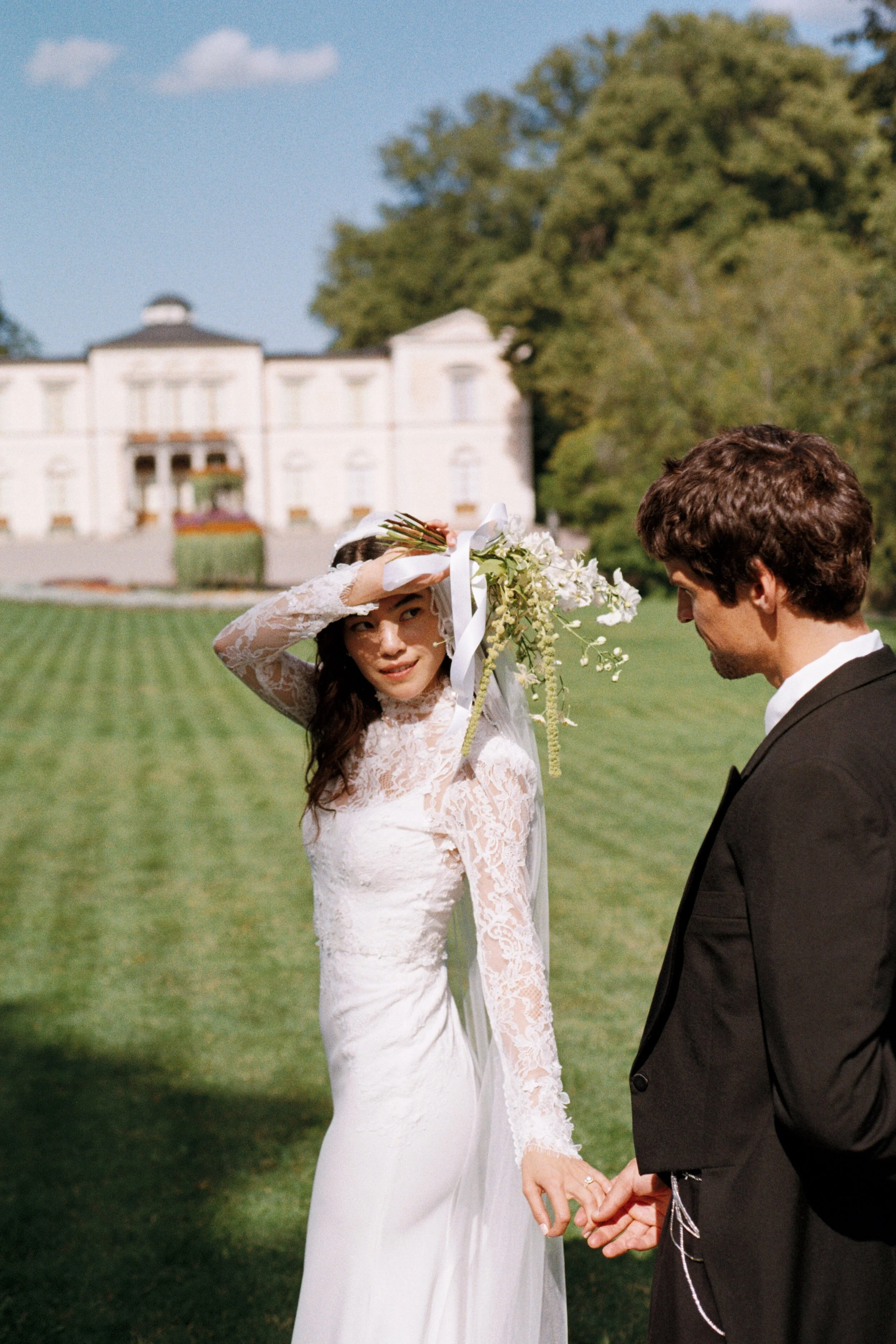 A bride in a white lace wedding dress holding a bouquet and shielding her eyes from the sun, standing outdoors with a groom in a black suit, in front of a large white mansion and green trees.