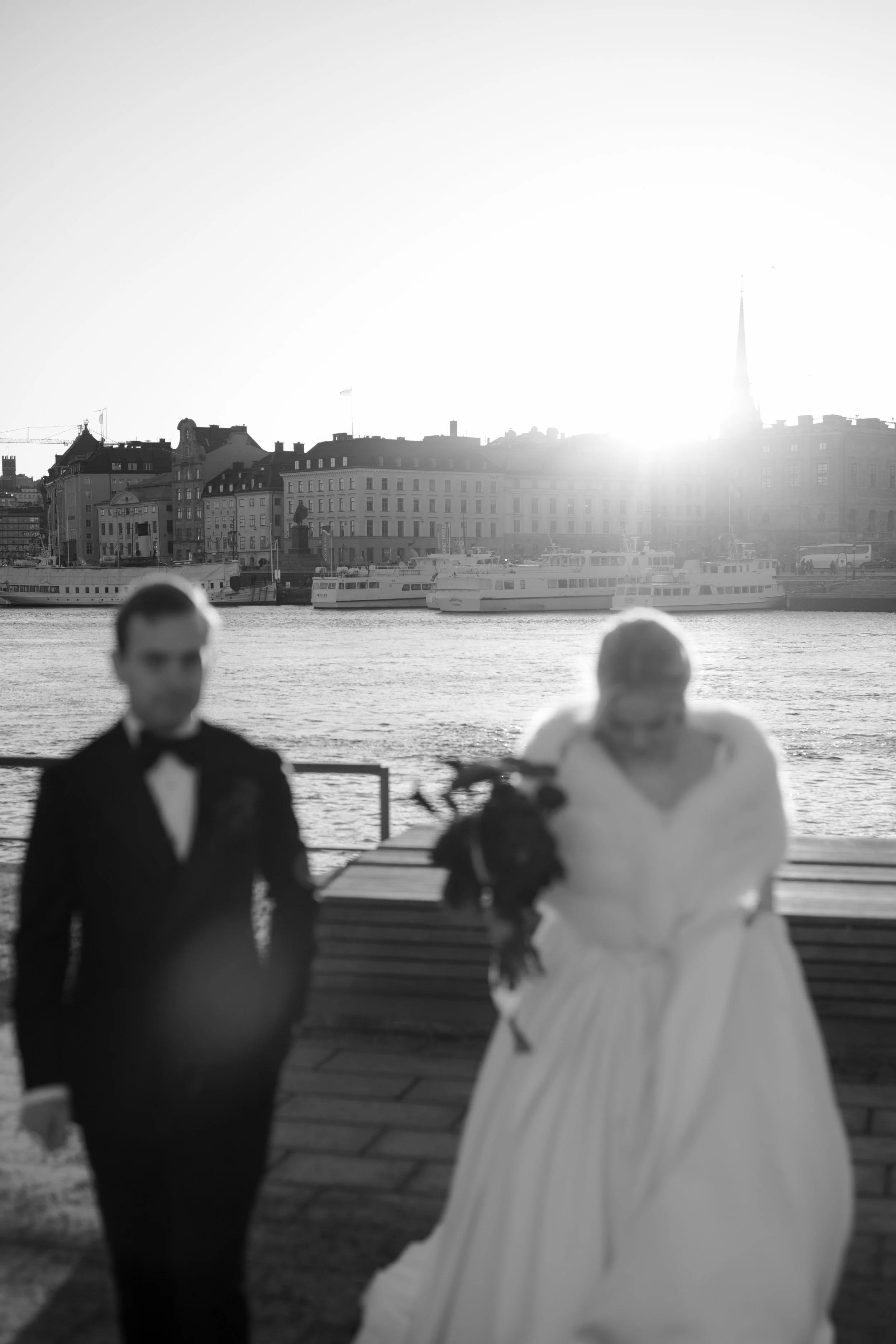 A black and white photo of a bride and groom walking by a waterfront with boats and city buildings in the background, sunlight shining brightly behind.