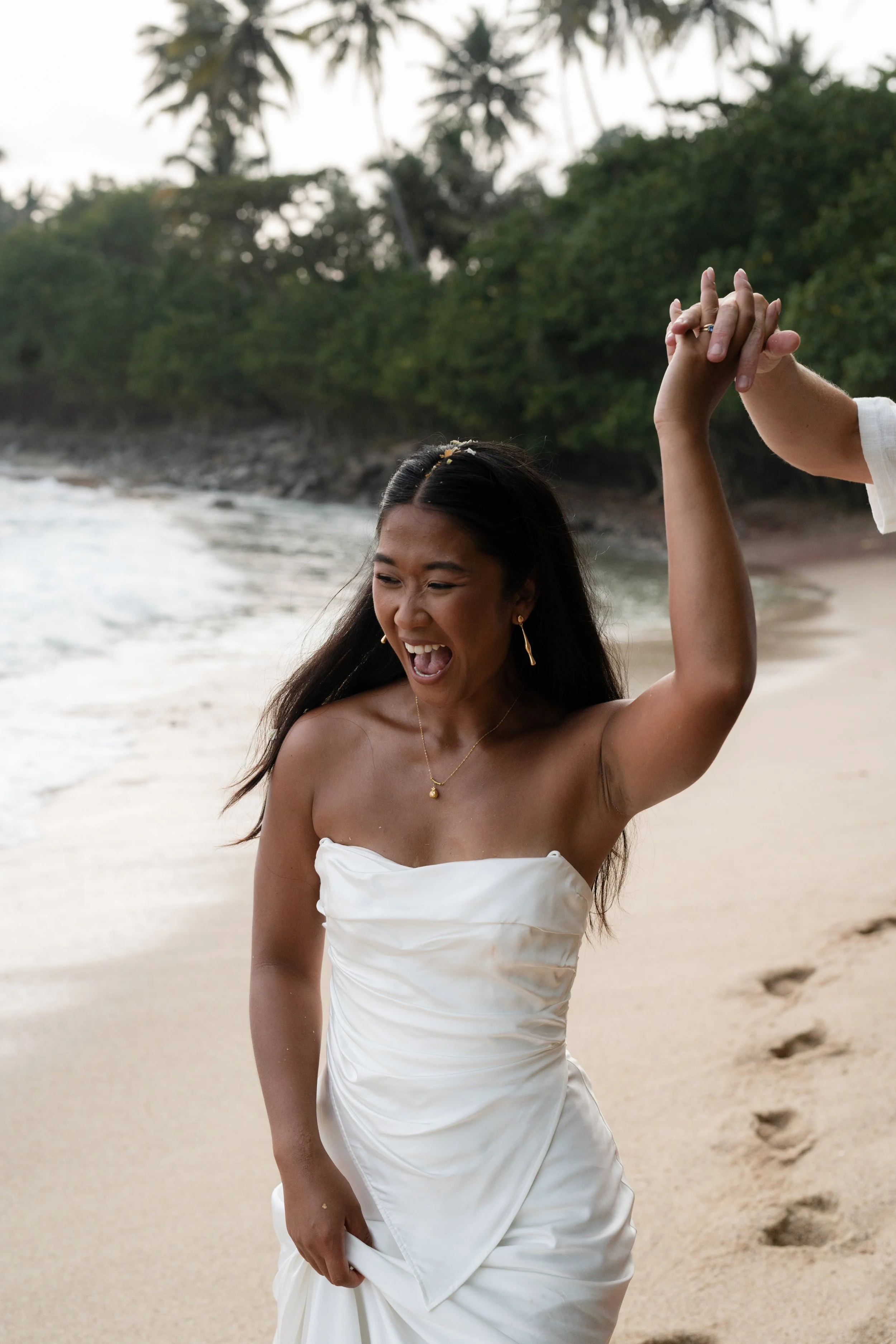 Woman in a wedding dress happily dancing on a beach, holding hands with someone off-camera, with footprints in the sand and palm trees in the background.