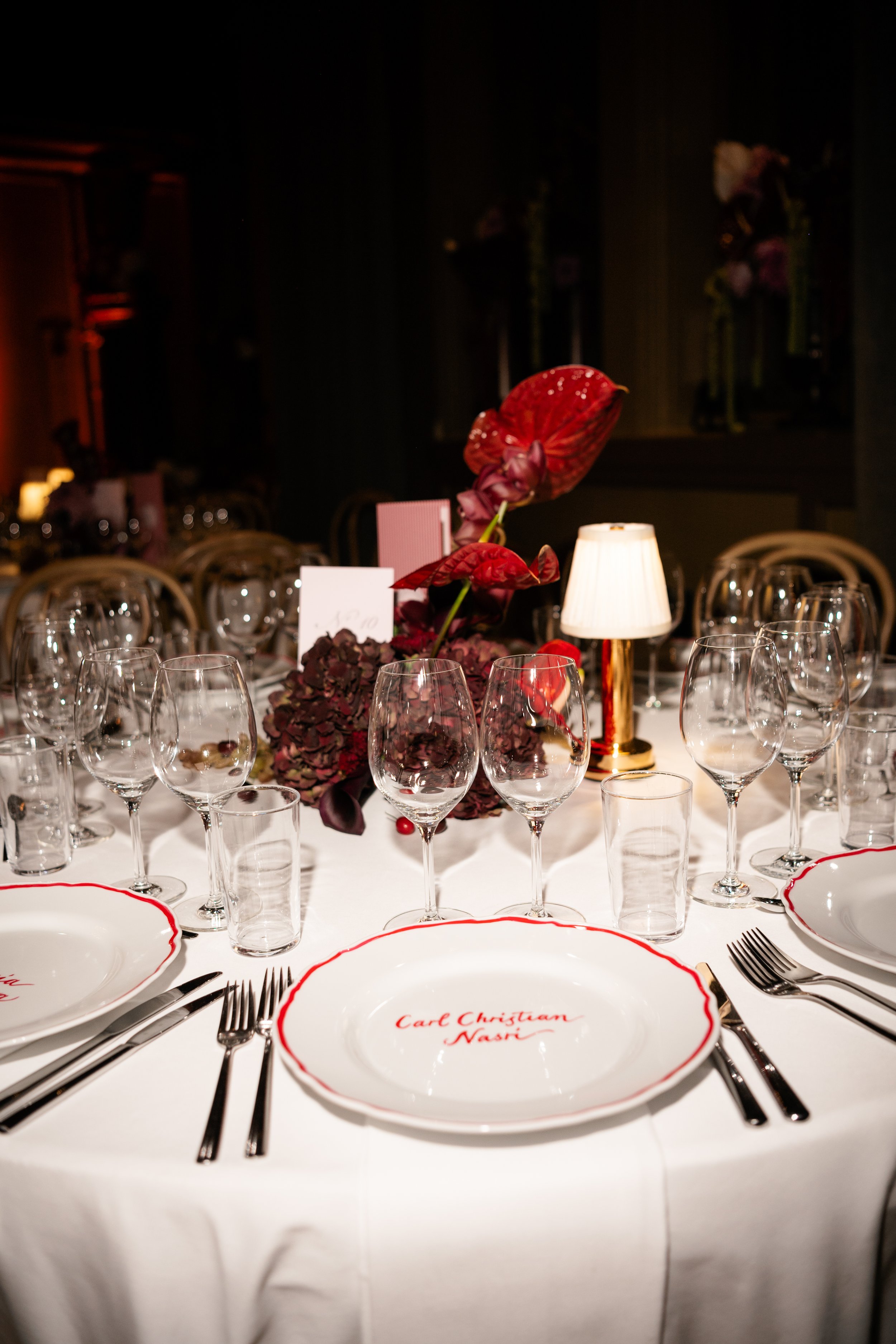 Elegant table setting with white plates, wine glasses, silverware, and a floral centerpiece with dark red flowers, illuminated by a small table lamp, at a formal event or wedding reception.