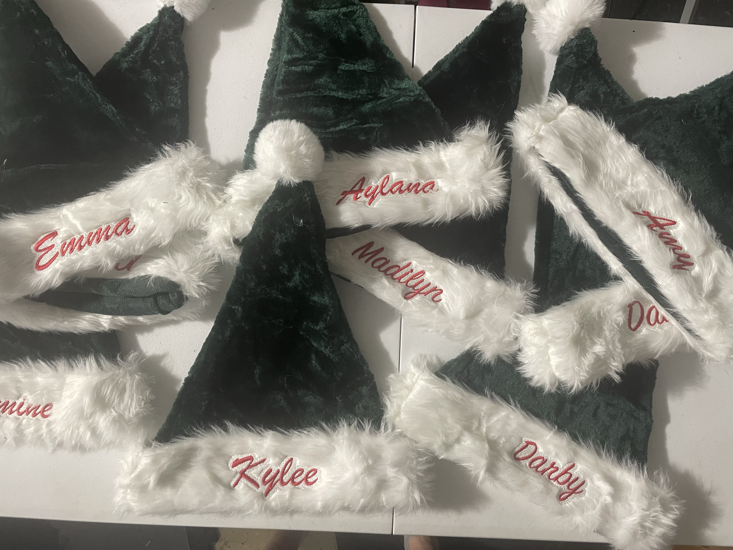 Several Christmas-themed hats and headbands with white faux fur trim and dark green velvet fabric, embroidered with red names including Emma, Aylane, Madilyn, Kylee, Daddy, and Daphney, laid out on a white surface.