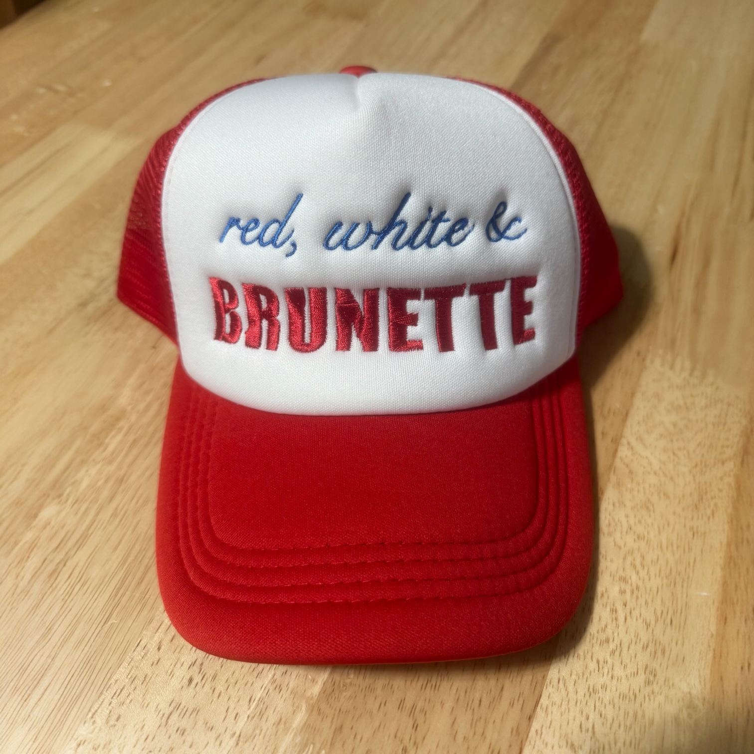 Red and white trucker hat with blue embroidered text saying 'red, white &' and red embroidered text saying 'BRUNETTE', on a wooden surface.