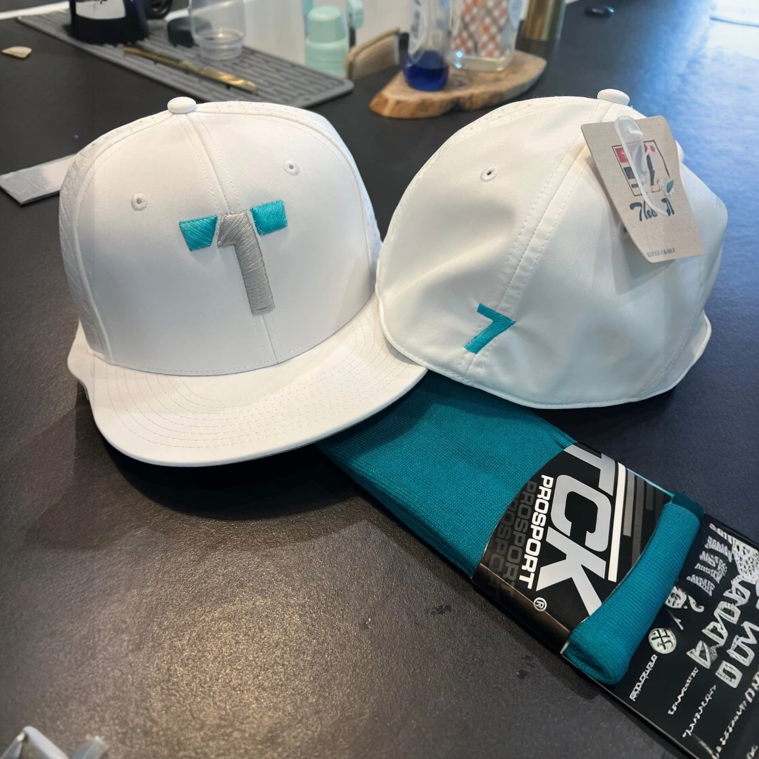 Two white baseball caps with embroidered logos, one showing the letter 'T' with teal and gray accents, and the other with a teal '7' on the side. A teal and black ProSport sock is also visible.
