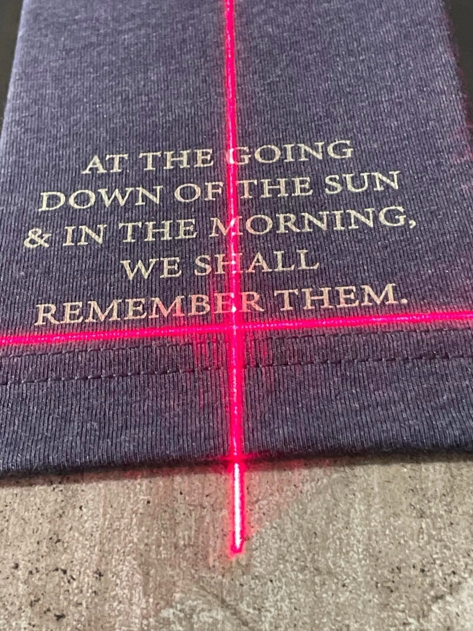 Text on a fabric surface reads, 'At the going down of the sun & in the morning, we shall remember them,' surrounded by a neon pink laser grid.