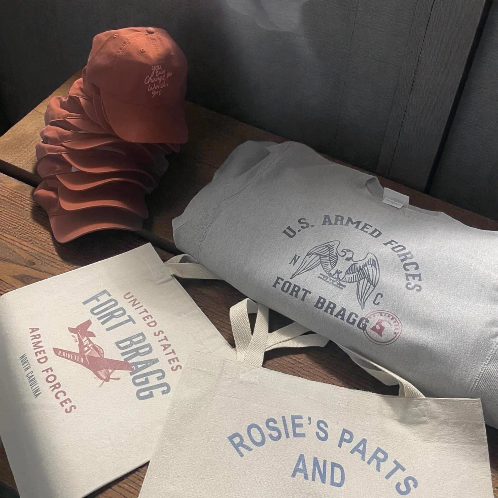 Stack of pink hats, a gray U.S. Army Fort Bragg sweatshirt, and two tote bags with vintage military aircraft and insignia designs on a wooden surface.