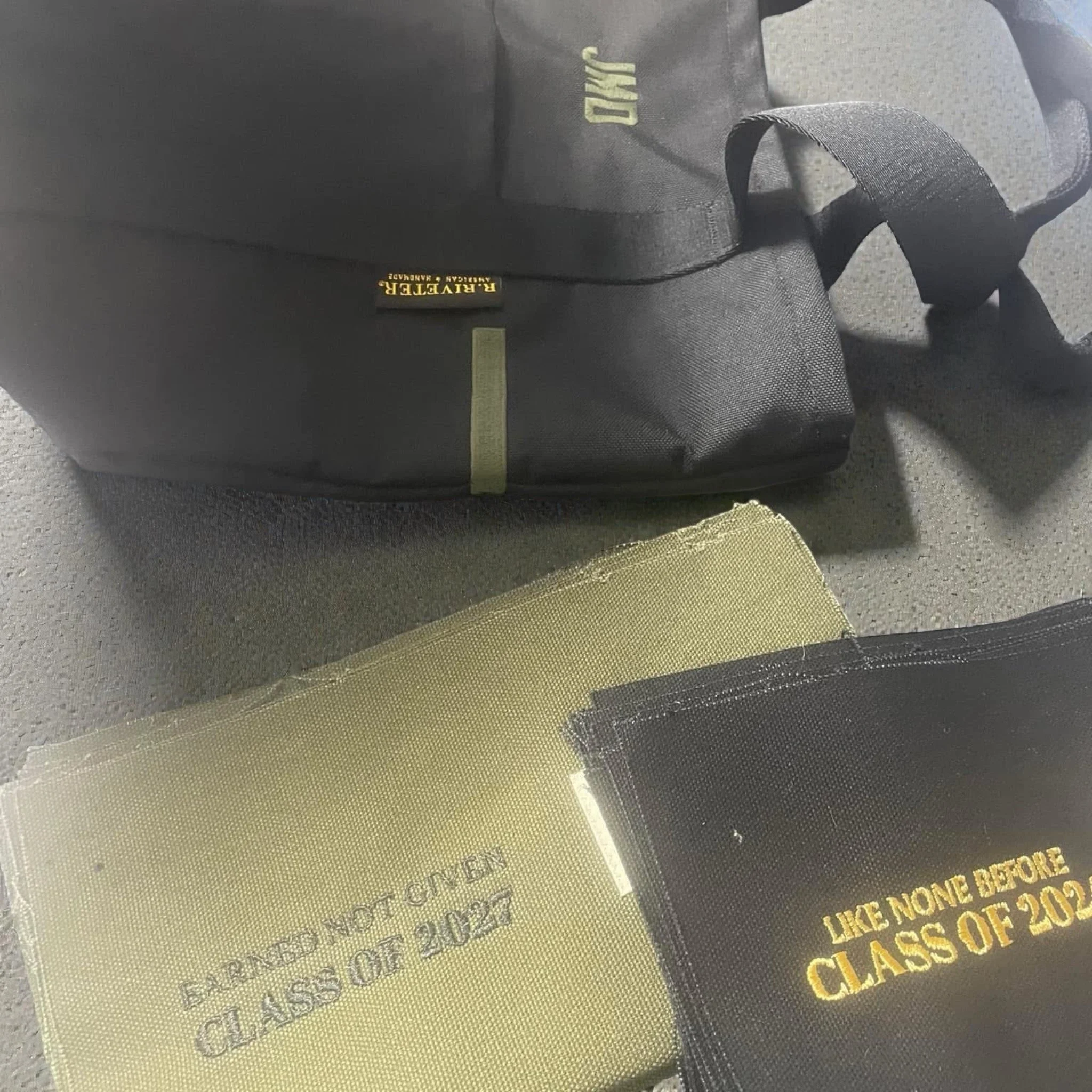A black RAVEETER backpack and two graduation programs for the class of 2022, one gold and one black, with the black one showing the text 'LIKE NONE BEFORE CLASS OF 2022' in gold lettering.