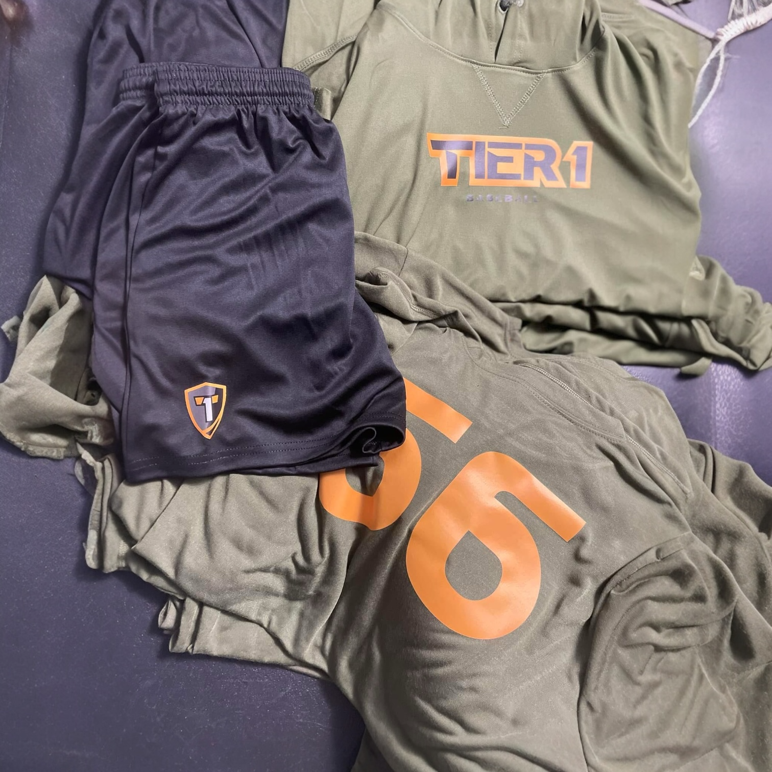Clothing items on a black surface, including a black pair of shorts with a small logo, gray sweatpants with a large orange number, and a green hoodie with a colorfully styled 'TIER 1' logo.