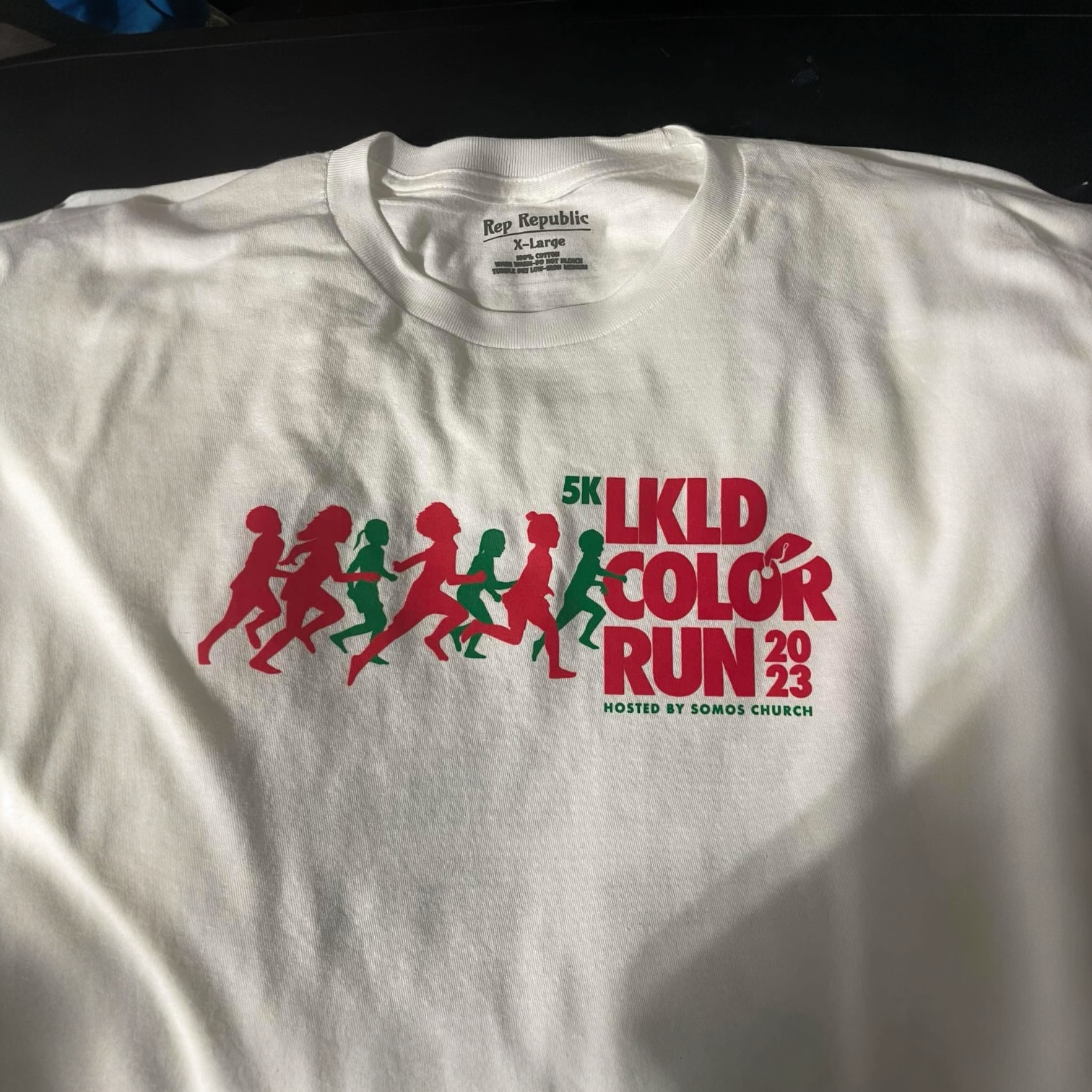 White T-shirt with a 5K run event design in red and green, featuring running silhouettes and text that reads 'LKLD COLOR RUN 2023' hosted by Somos Church.