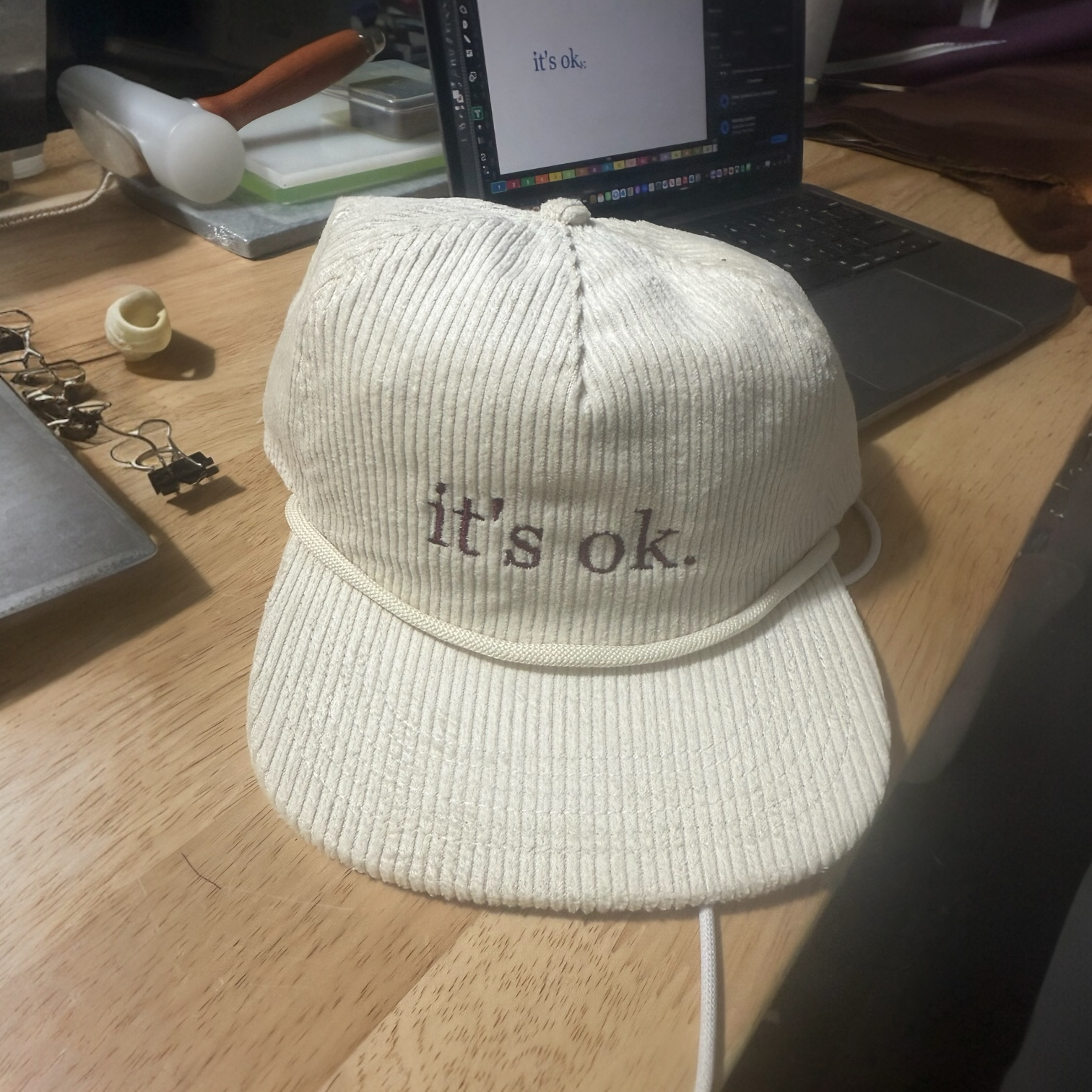 A beige corduroy baseball cap with the words "it's ok." embroidered on the front, placed on a wooden table in front of a laptop.