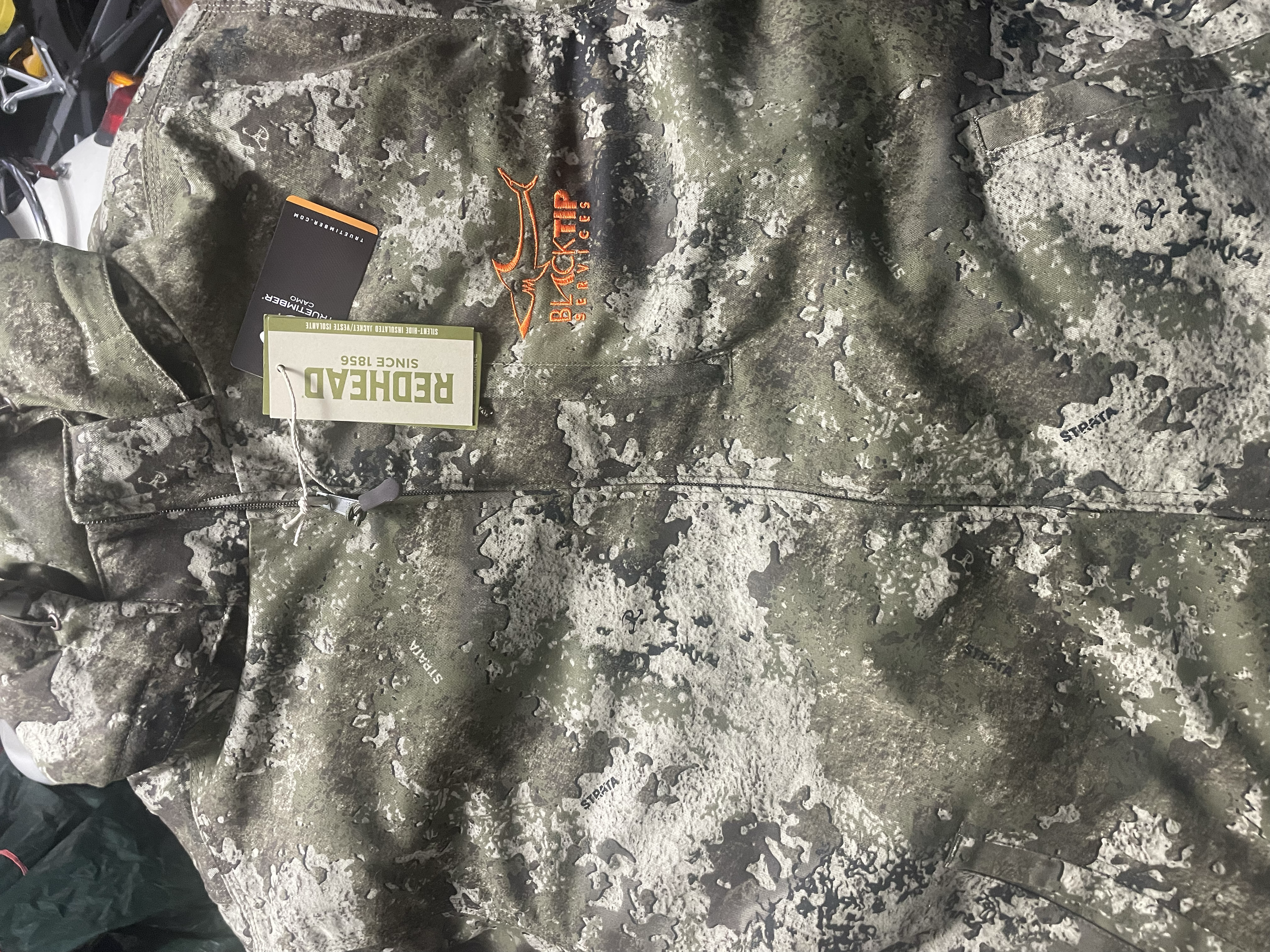 Close-up of a camouflage patterned jacket with an orange embroidered logo of a fish and the words "Blacktip Services." The jacket has a brand tag "Reohard" and a tag with the text "since 1856."