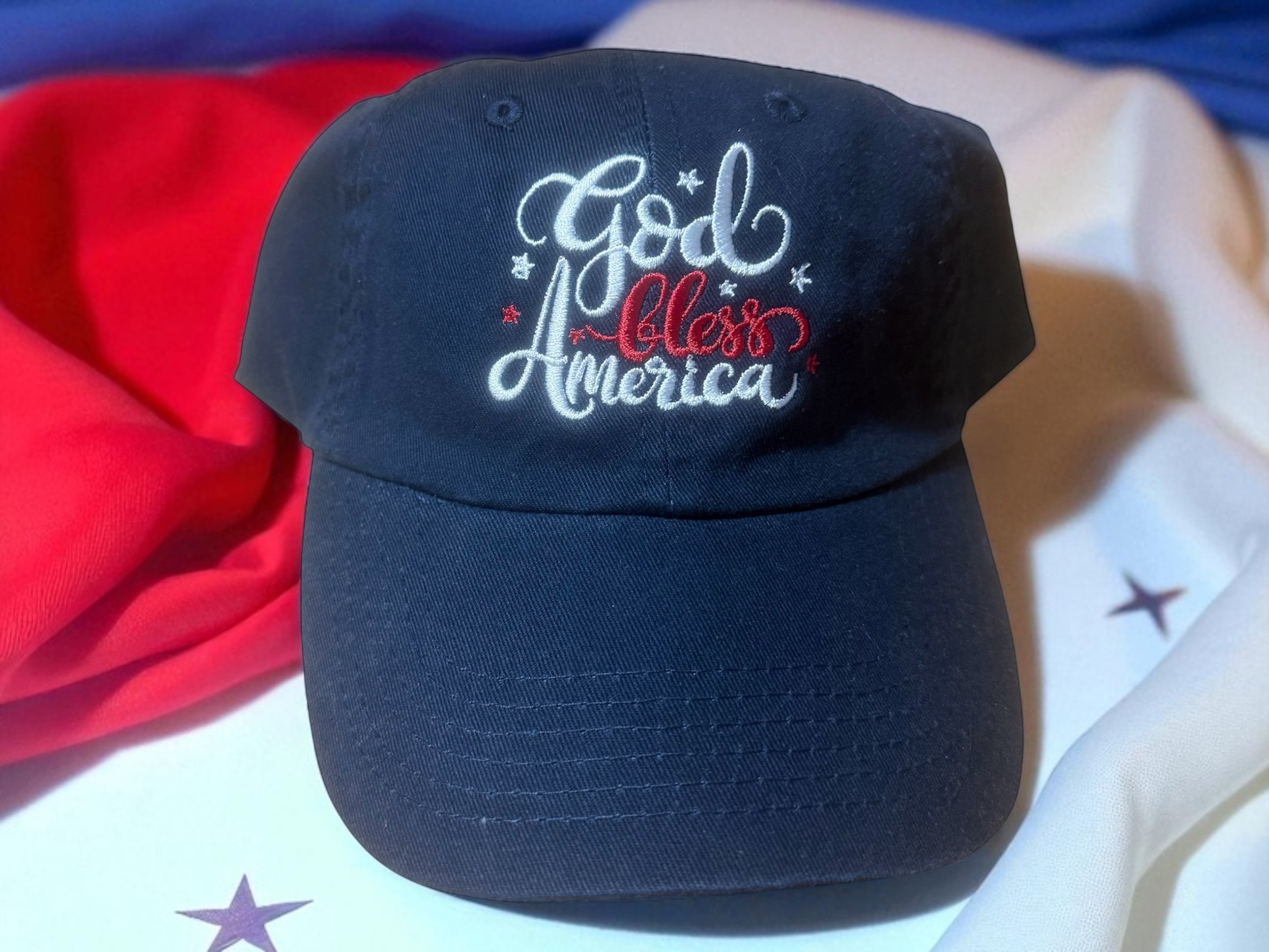 A navy blue baseball cap with the words "God bless America" embroidered on the front, surrounded by small stars. The cap is placed on a surface with red, white, and blue fabric.