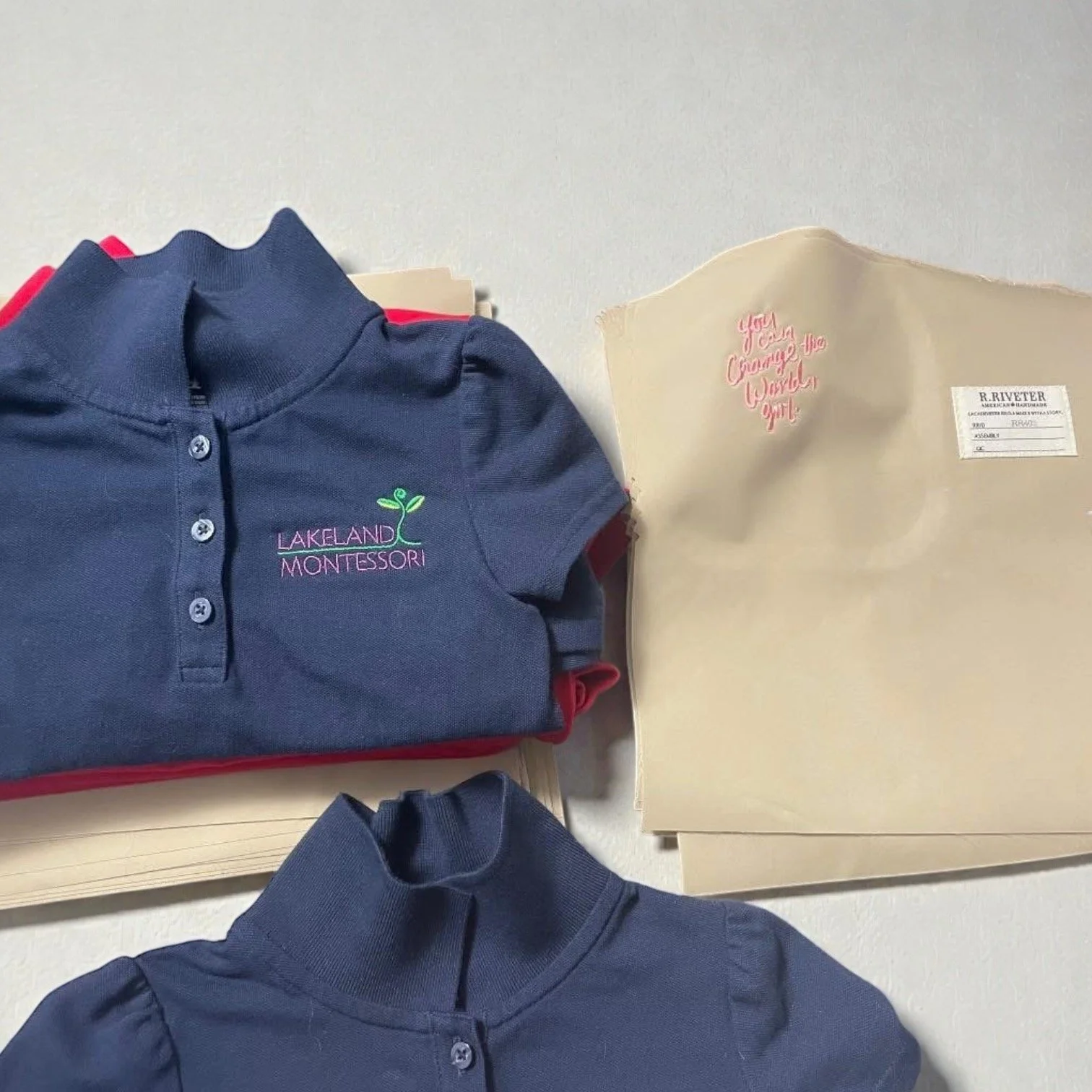 School uniforms and folders on a white surface, embroidered with 'Lakelands Montessori' and 'You Can Change the World Girl'.