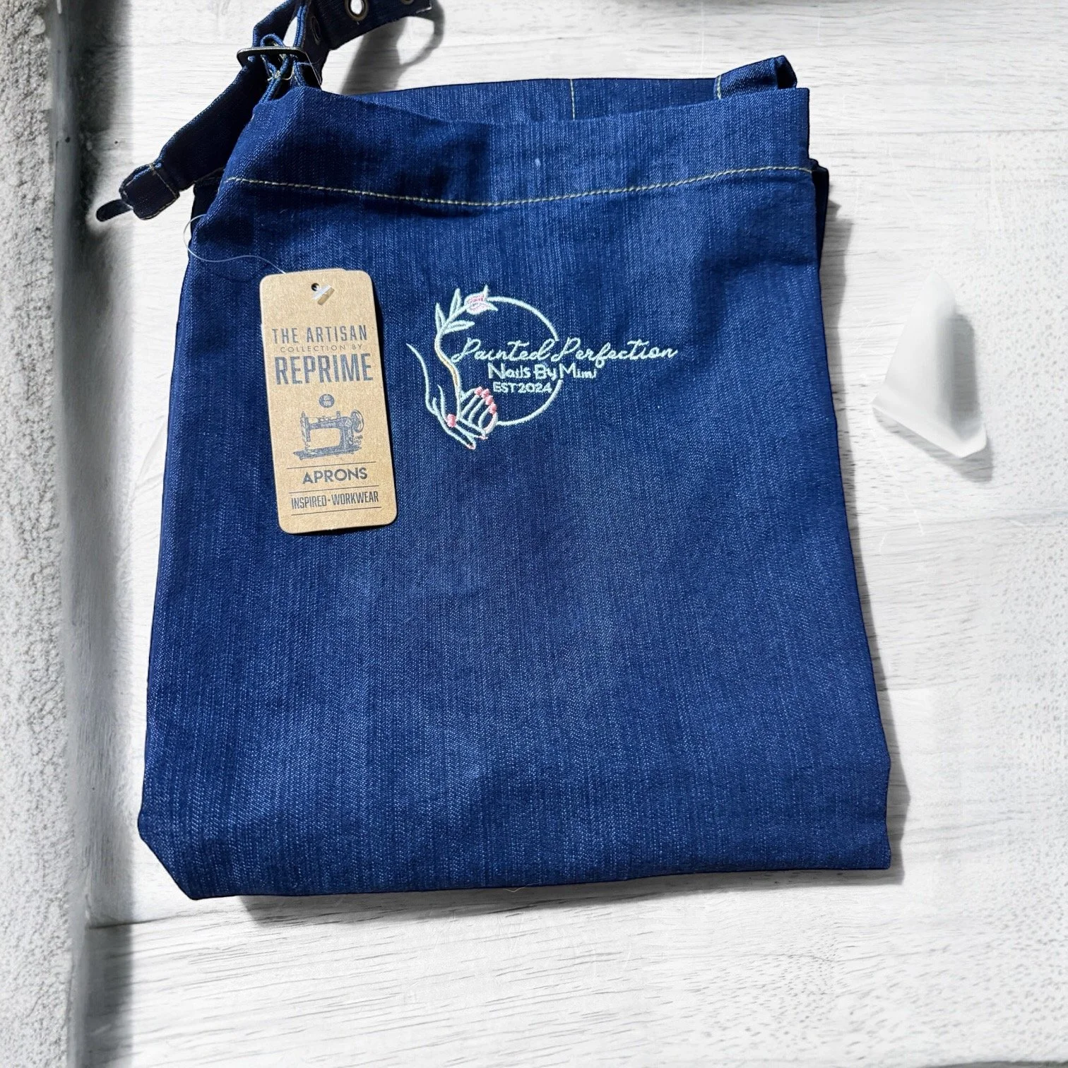 Folded blue denim apron with embroidery and a cardboard tag attached, resting on a white surface near a wall.