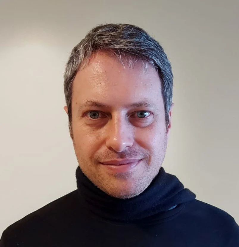 Retrato de um homem com cabelo castanho grisalho, pele clara, usando suéter preto, sorrindo e olhando para a câmera.