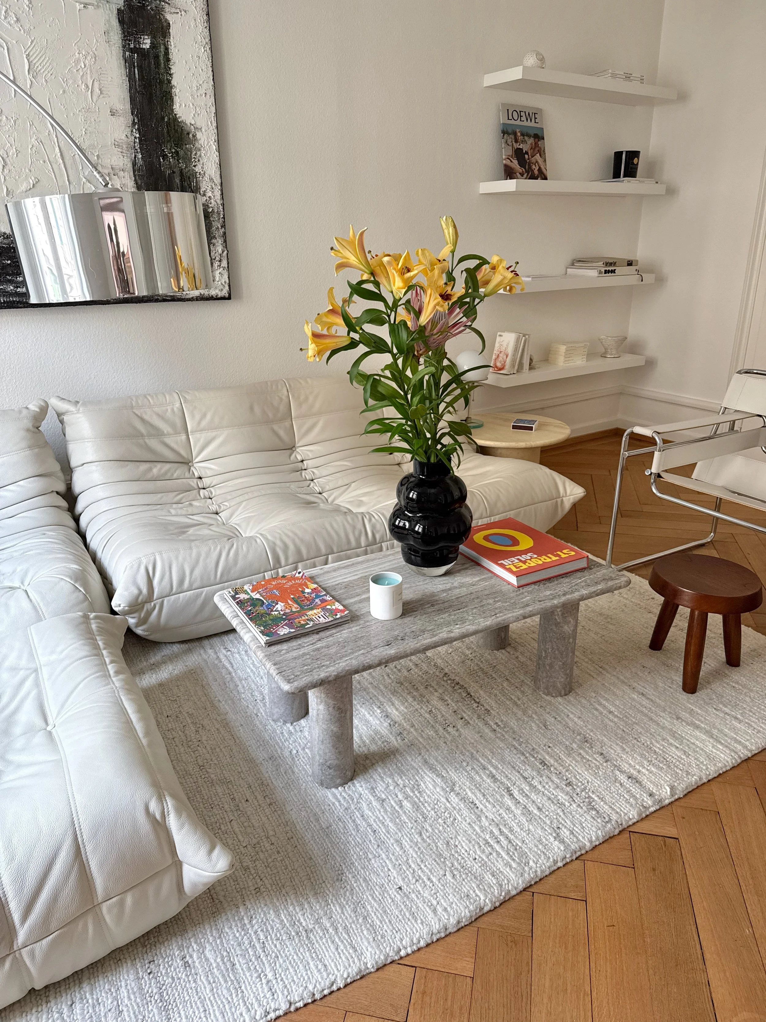Living room with a white tufted sofa, a rustic stone coffee table with books and a small candleholder, a large black vase with yellow lilies, a wall with white textured paint, a painting, and wall shelves with books and decorative items. There's a white armchair with chrome frame and a small wooden stool.