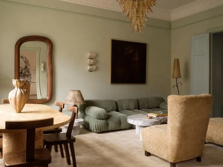 Living room with a green sofa, a beige armchair, a wooden round dining table with chairs, a mirror on the wall, a floor lamp, and decorative wall art.