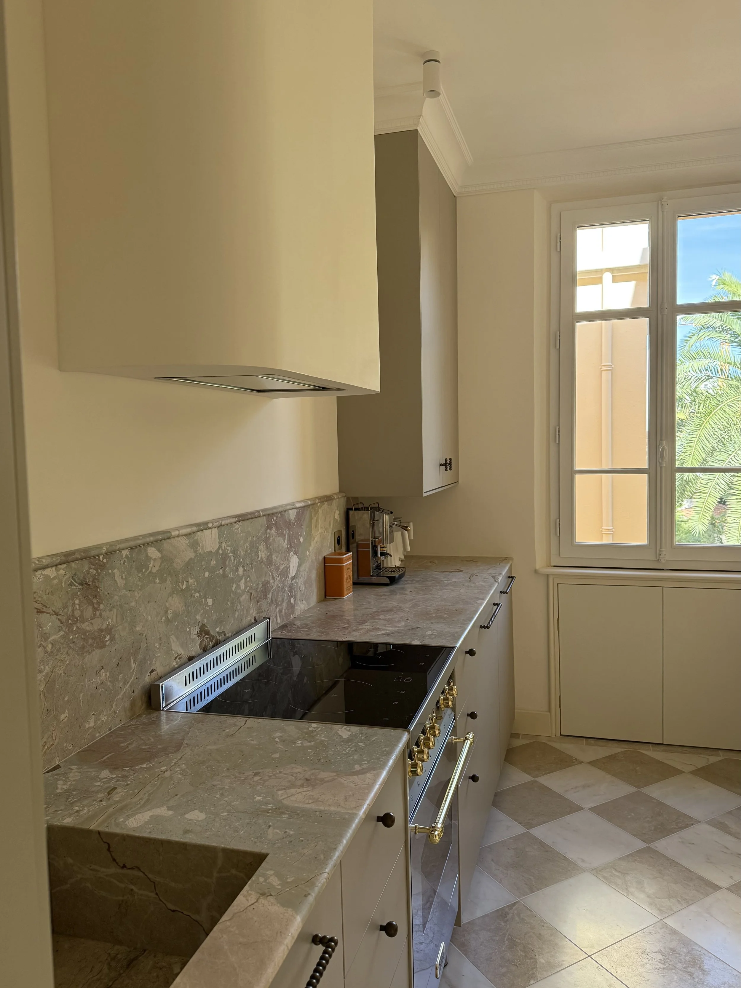 A kitchen with a marble countertop, beige cabinets, a window with a view of trees, and a stove with gold knobs.