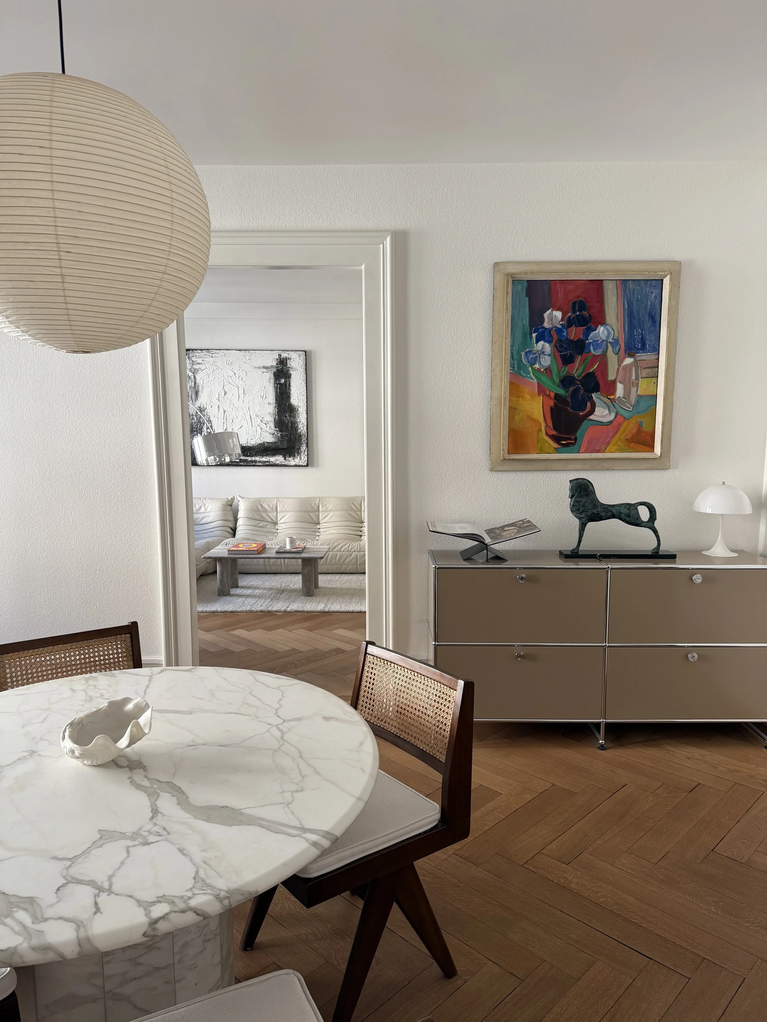 An interior view of a modern living room and dining area with white walls, wooden flooring, and contemporary furniture. There is a marble dining table with wooden chairs, a beige cabinet with decorative items, and colorful artwork on the walls.