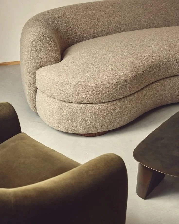 A beige, textured, modern sofa with rounded edges, next to a dark wood coffee table and a brown armchair, on a light-colored floor.