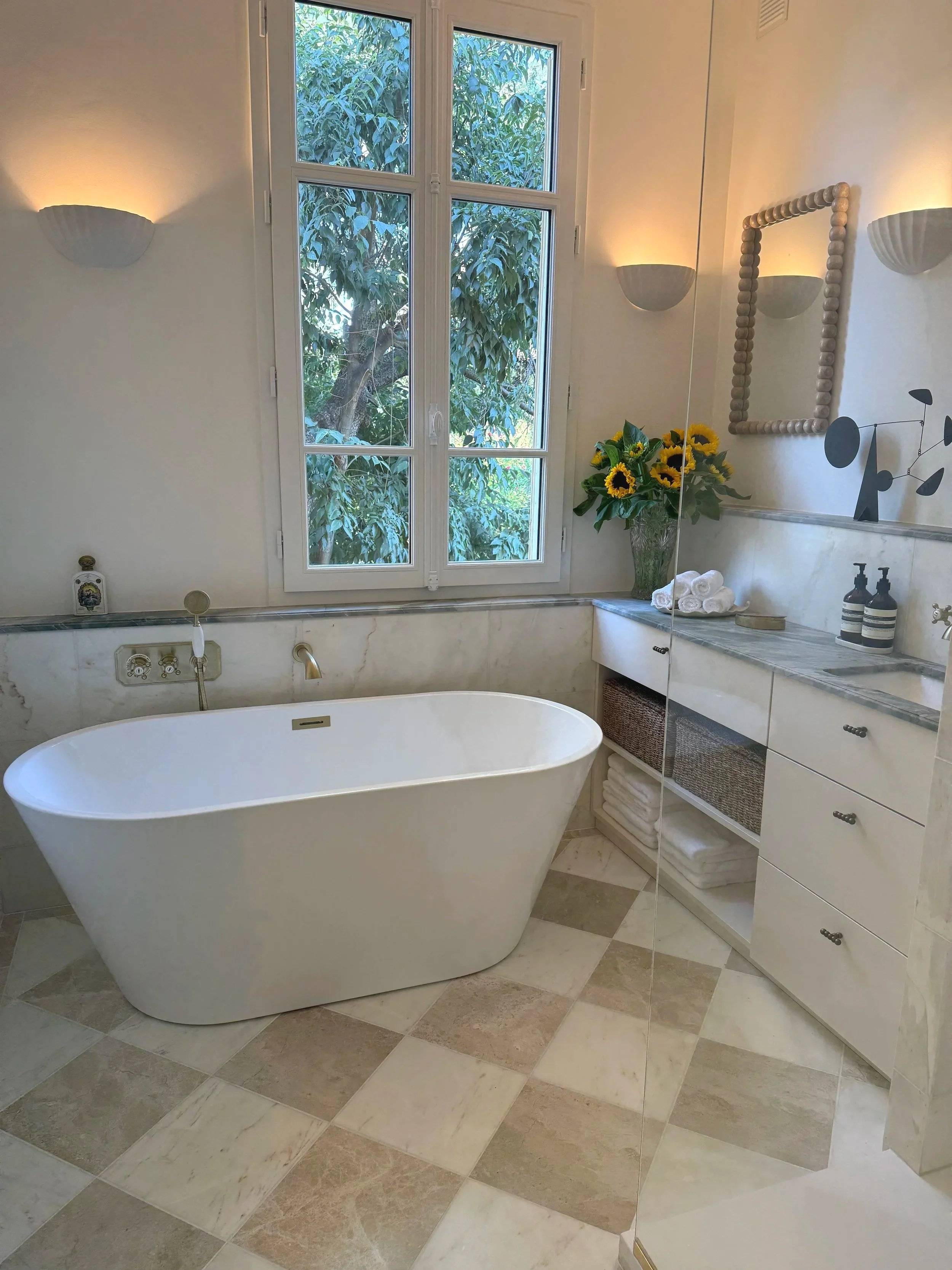 A bathroom with a white freestanding bathtub positioned near a large window with views of green trees outside. The bathroom has beige and off-white tiled floors, a marble countertop with rolled white towels, a vase with sunflowers, and a mirror with a bead frame. Wall-mounted sconces provide lighting.