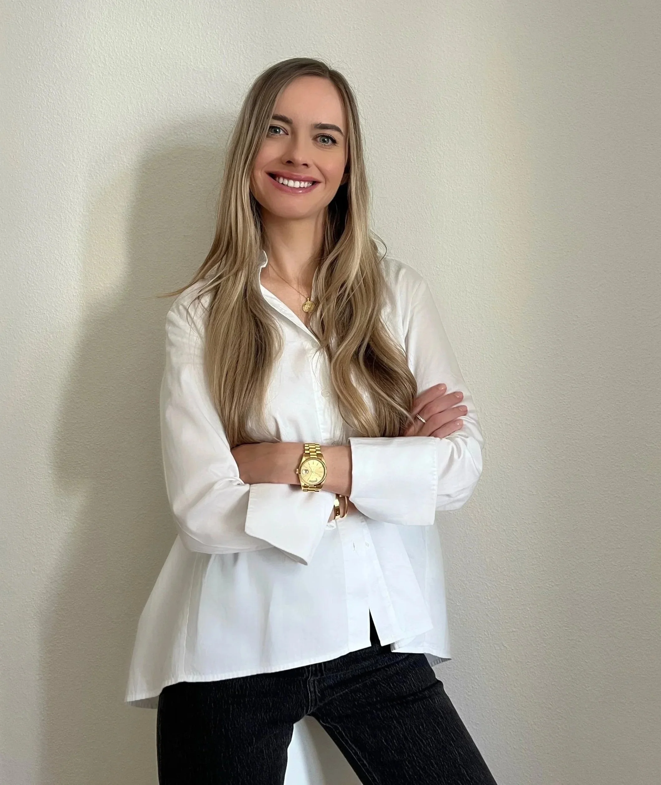 A woman with long blonde hair wearing a white shirt, black pants, a gold watch, and a gold necklace, standing with her arms crossed against a plain beige wall.