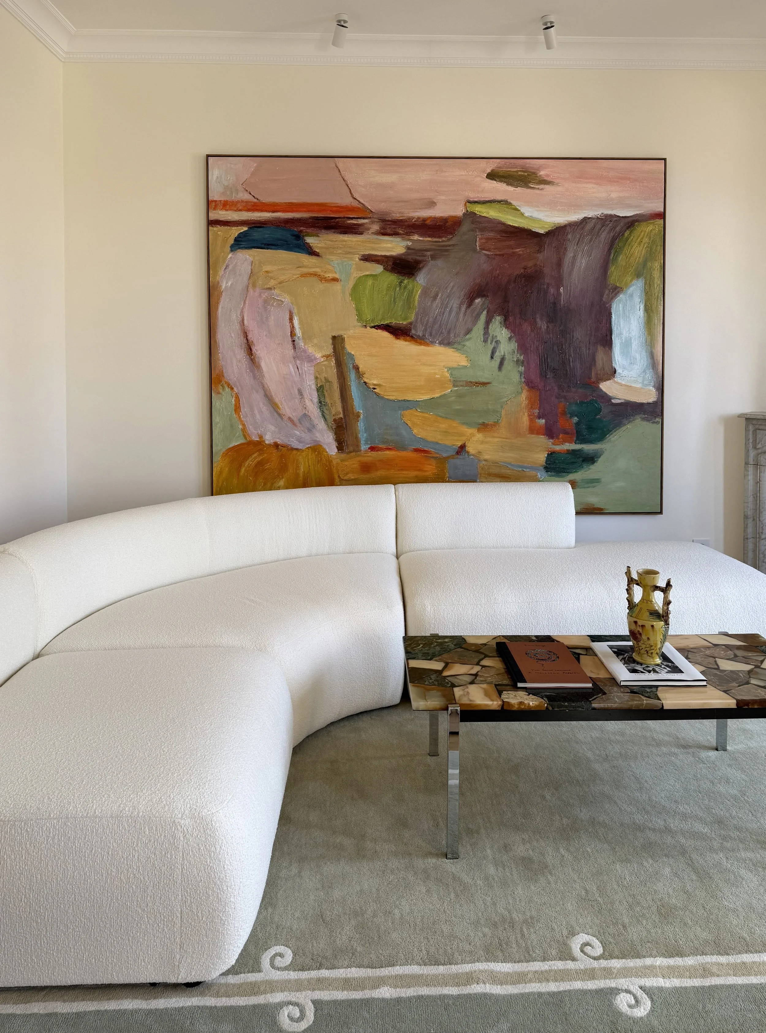 Living room with a curved white sofa, a marble coffee table with books and a decorative vase, a large abstract painting on the wall, a gray carpet with a white border, and a small section of a marble fireplace visible.