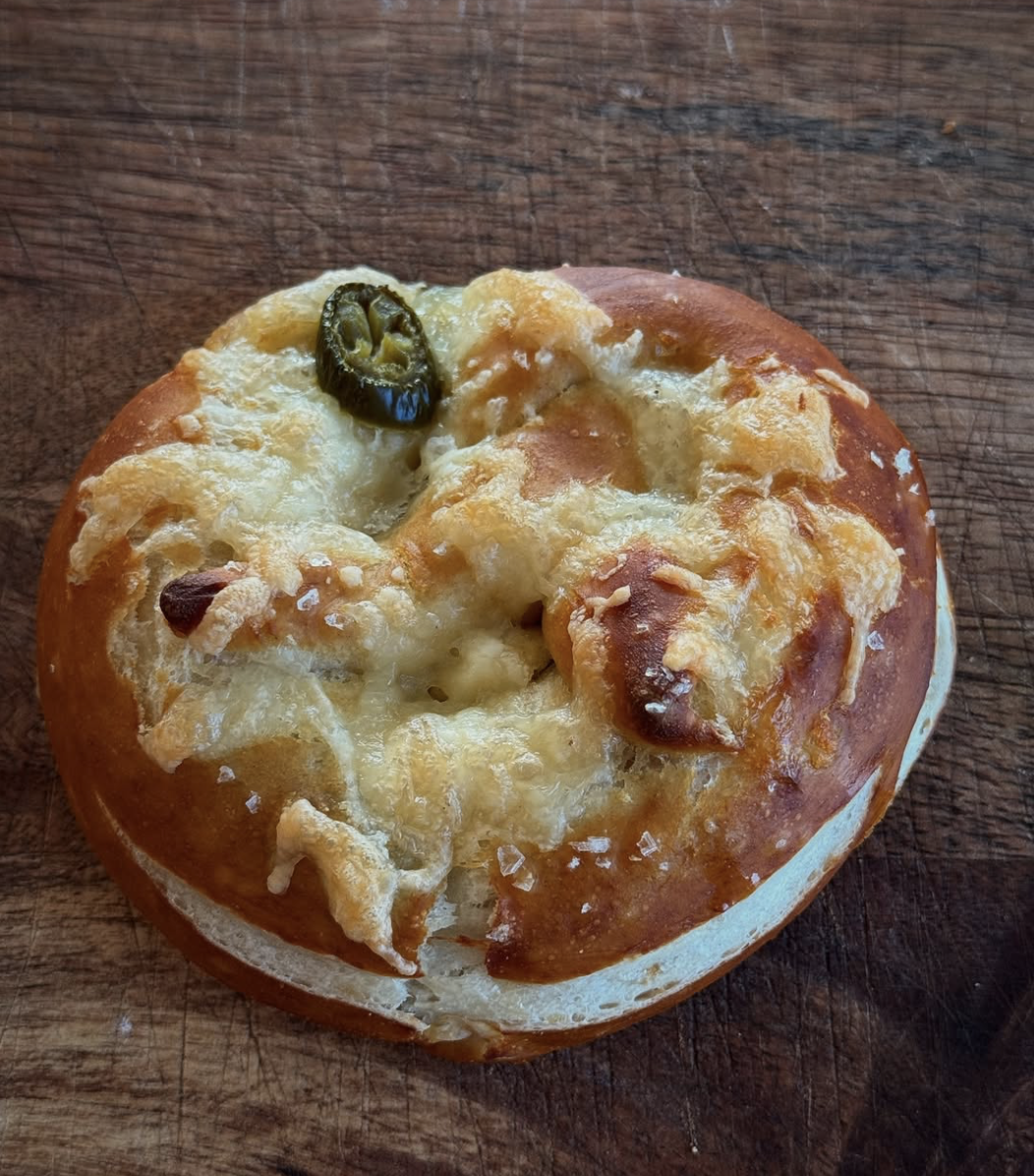 A round bread roll topped with melted cheese, a black olive, and bits of additional cheese on a wooden surface.