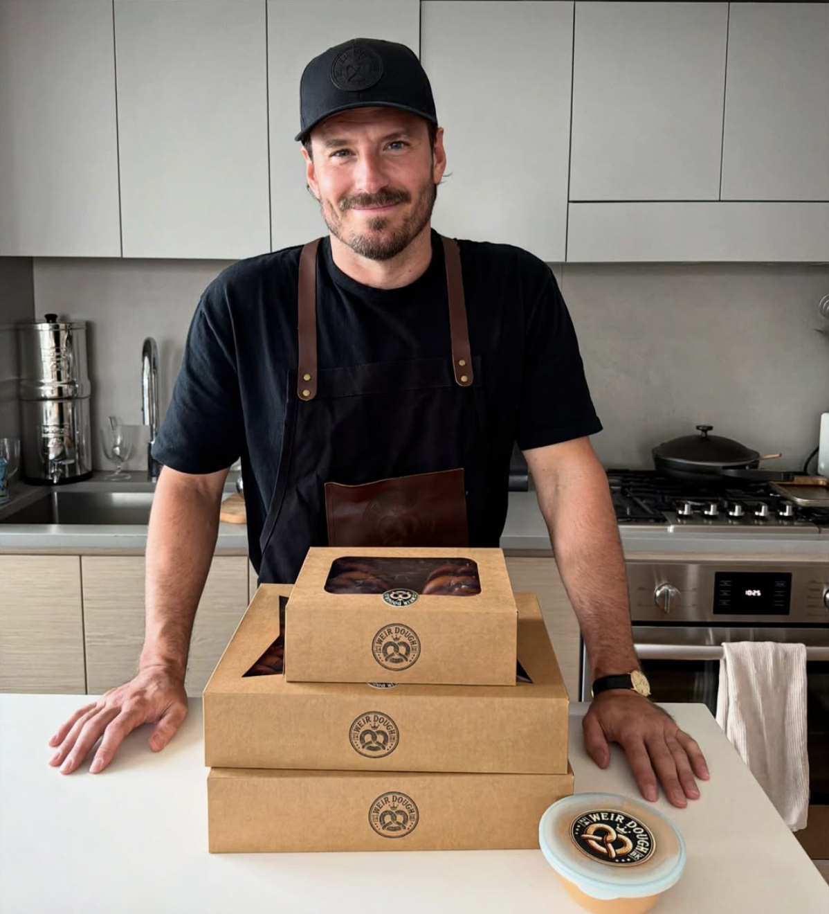 A man wearing a black cap and black apron stands behind a white kitchen counter, leaning on it with both hands. In front of him are three stacked cardboard boxes with a bakery logo, and a cake container with a logo is on the counter. The background features a modern kitchen with a stove, oven, and various kitchen items.