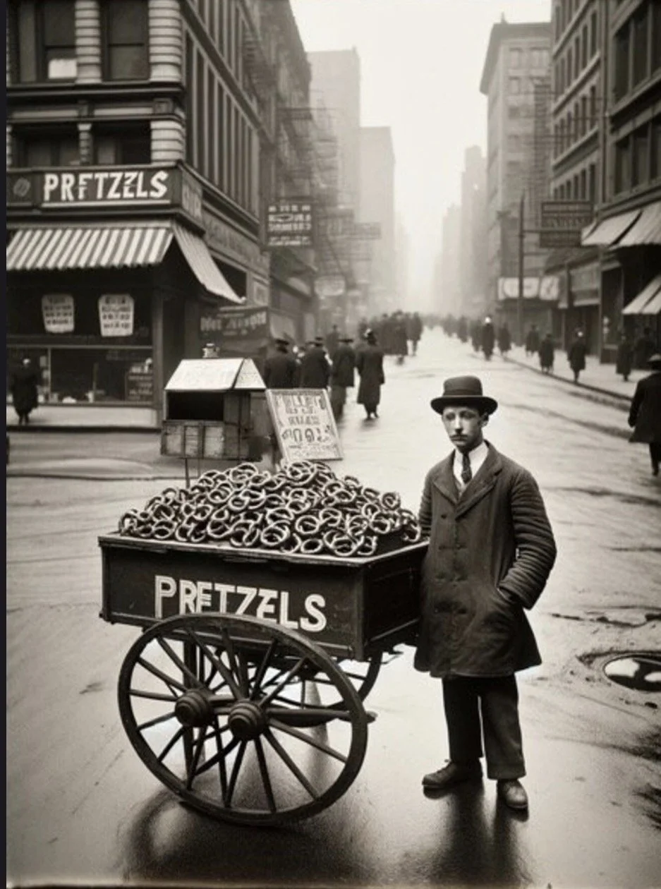 A man in a coat and hat standing next to a cart filled with pretzels on a city street in an early 20th-century black and white photograph.