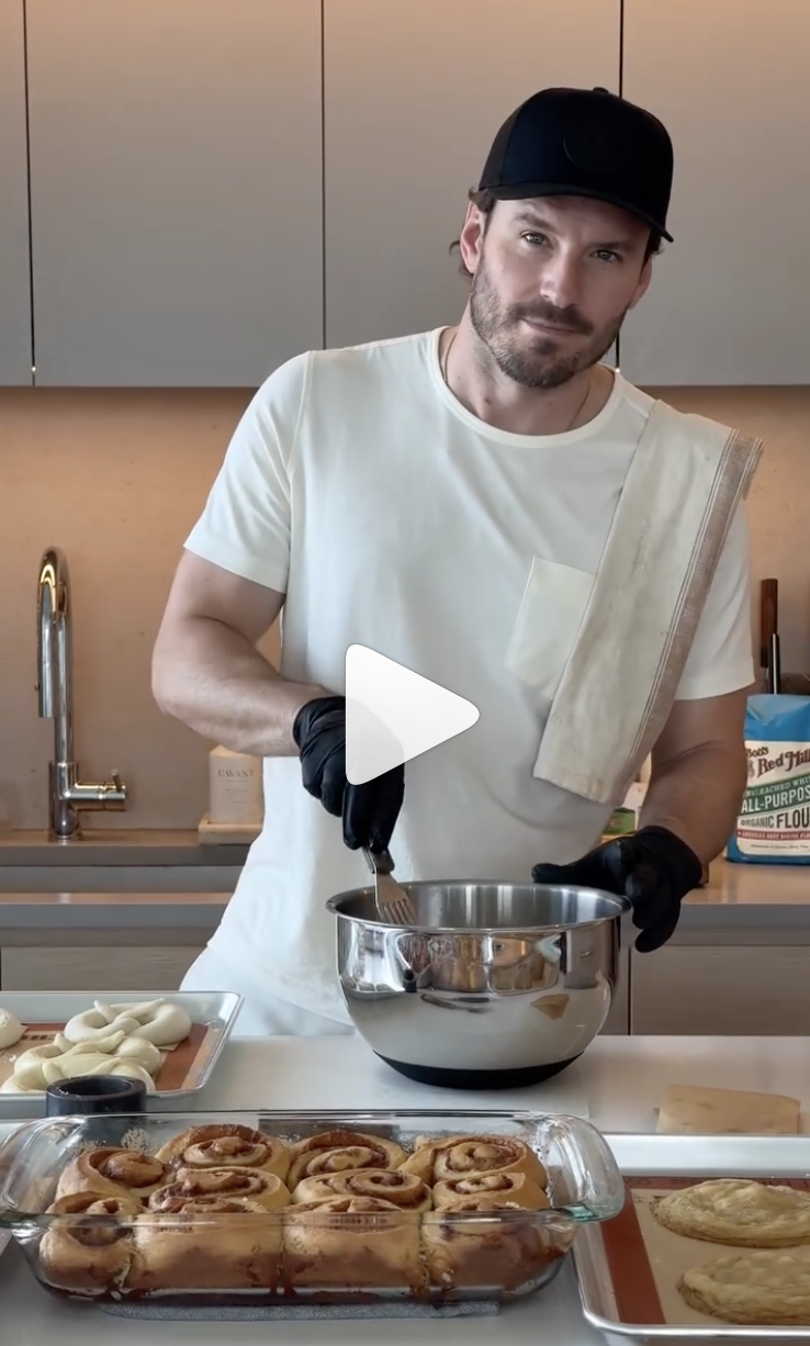 A man wearing a black cap, white t-shirt, and black gloves stands in a modern kitchen, holding a fork and working with a mixing bowl. There are cinnamon rolls in a glass baking dish on the counter in front of him, along with some bread or pastry dough on a tray, and a plate of white dough or frosting. The background features a sleek kitchen with a gray cabinet and a gold-colored faucet.