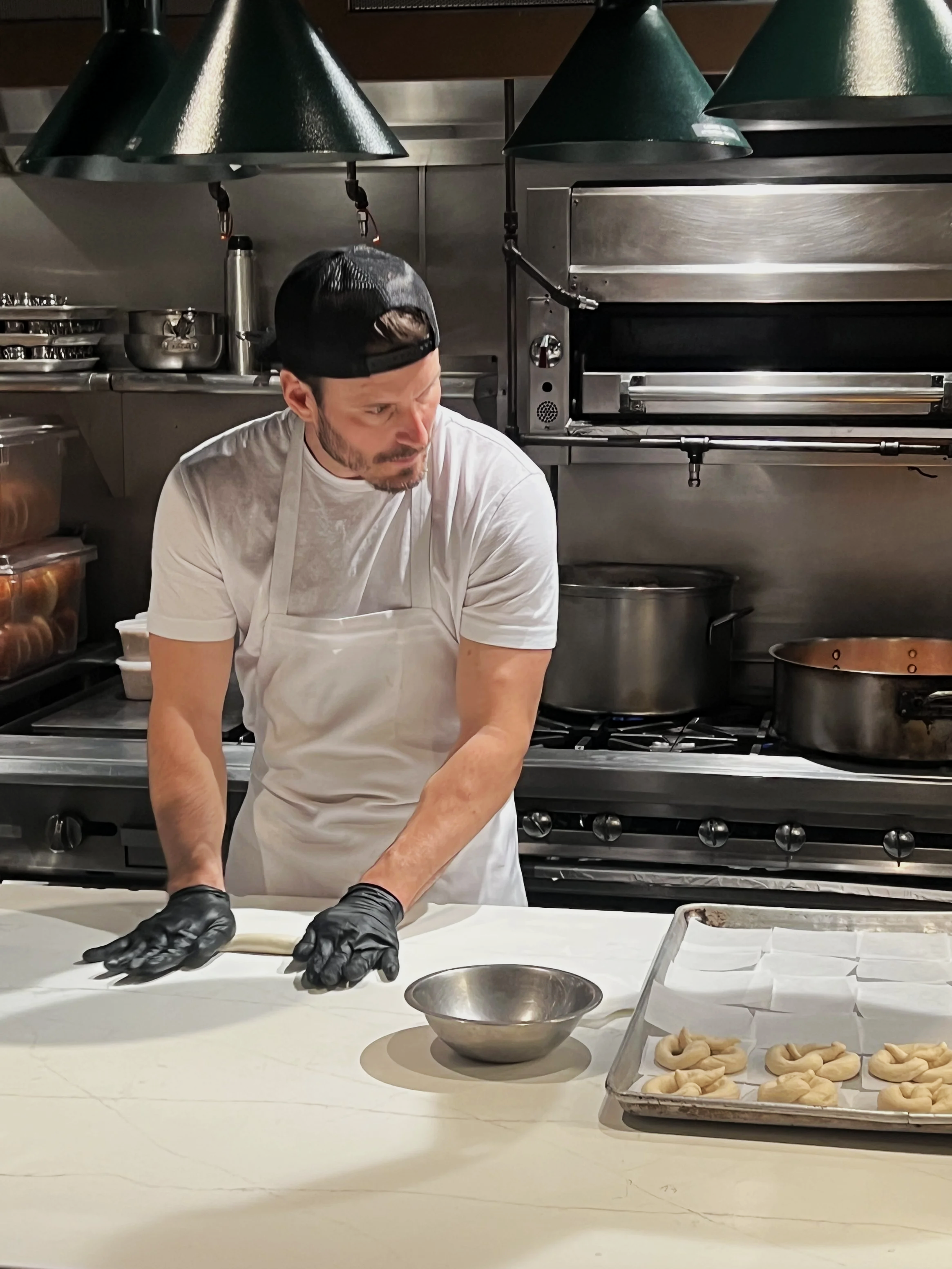 A man wearing a black backwards cap, a white t-shirt, a white apron, and black gloves is preparing dough in a commercial kitchen. There is a tray of shaped dough pieces on a baking sheet and a metal bowl on the counter in front of him. The background includes stainless steel appliances and kitchen equipment.